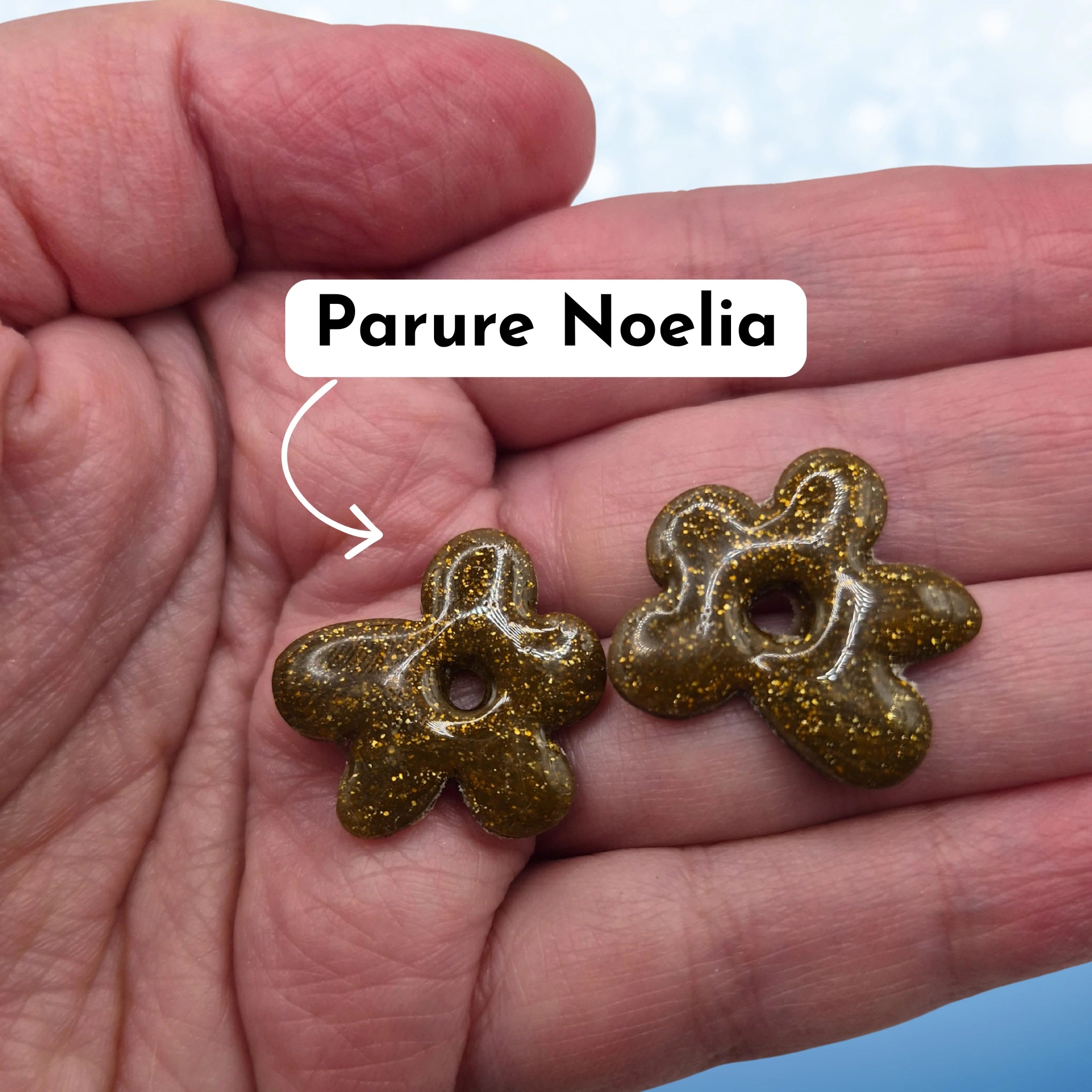 Pampilles en pâte polymère marrons, en forme de fleur difforme, avec l’inscription « Parure Noelia », présentées tenues dans une main, sur un fond en dégradé de bleu.