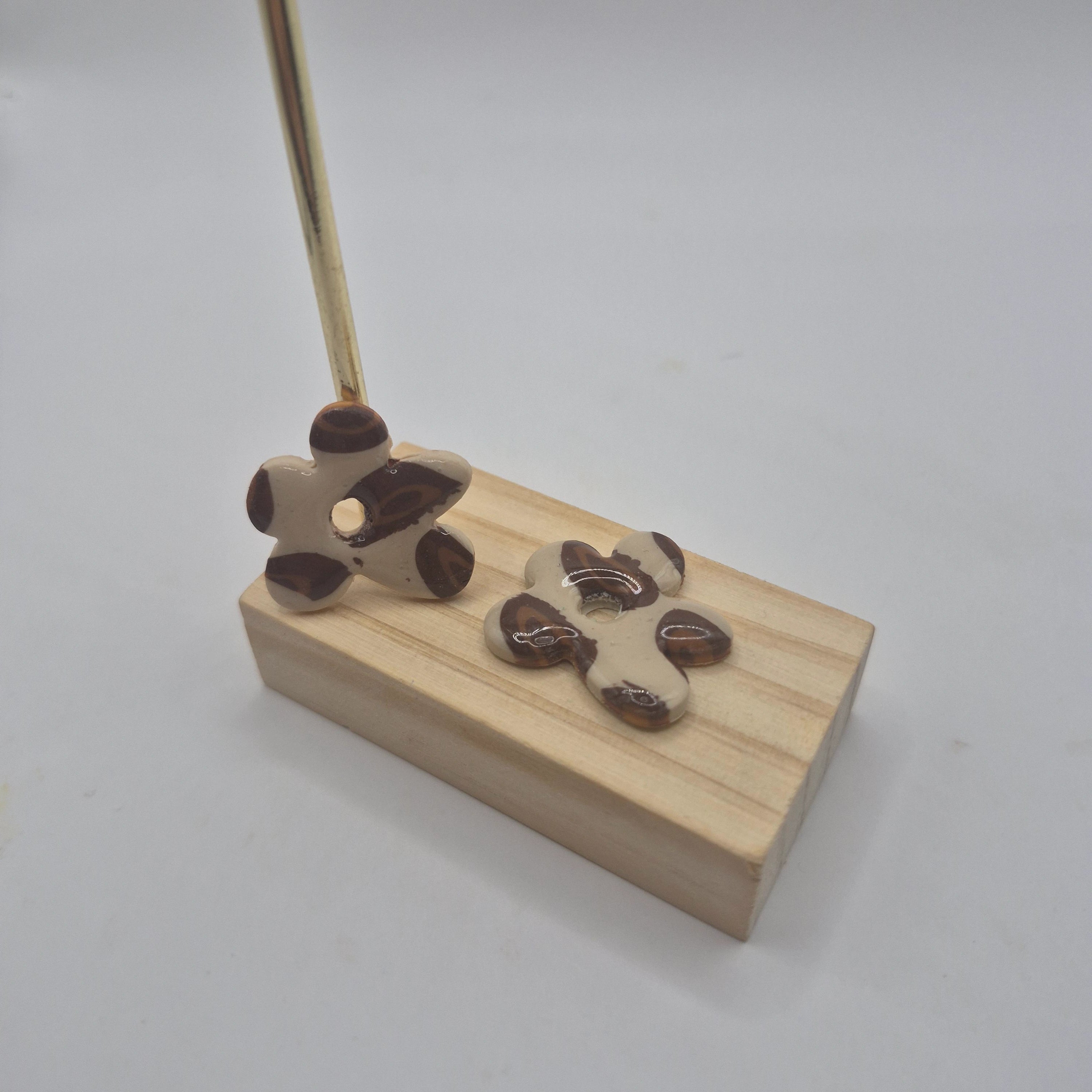 Boucles d’oreilles pampilles en forme de fleur irrégulière, en pâte polymère motif léopard beige et marron, faites main avec finition résine, présentées sur un support en bois aux côtés de deux créoles argentée — parure artisanale Noelia.