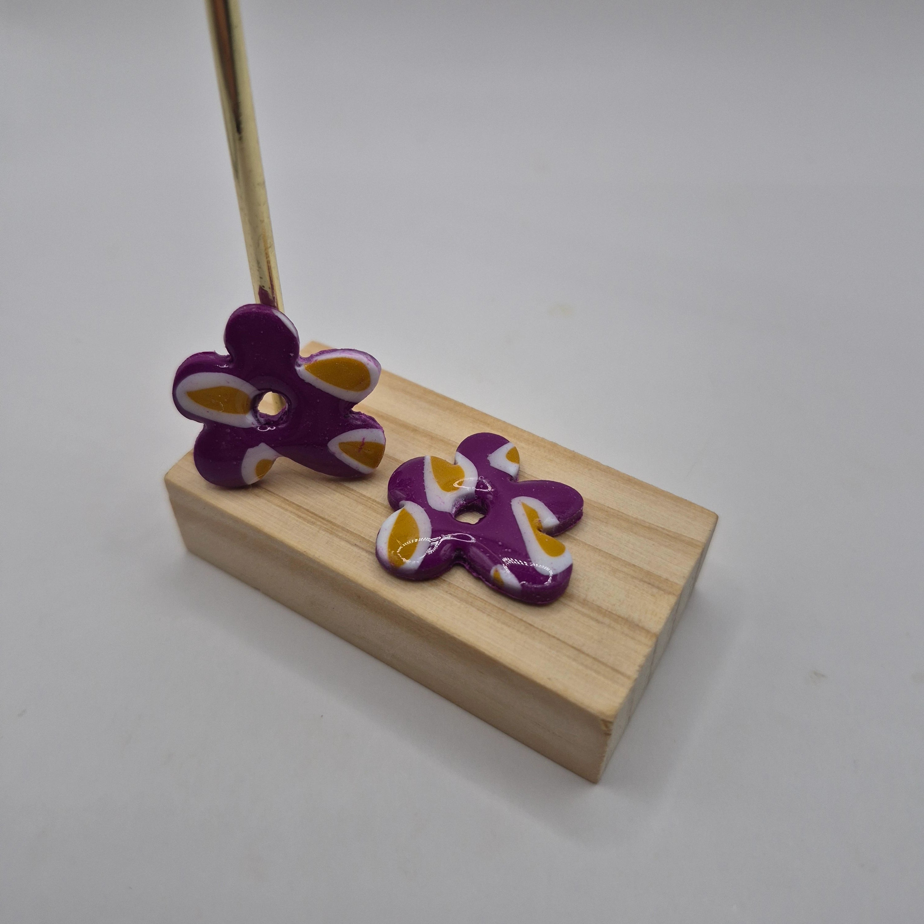 Boucles d’oreilles pampilles en forme de fleur irrégulière, en pâte polymère  violette, blanc et ocre, faites main avec finition résine, présentées sur un support en bois  — parure artisanale Noelia.