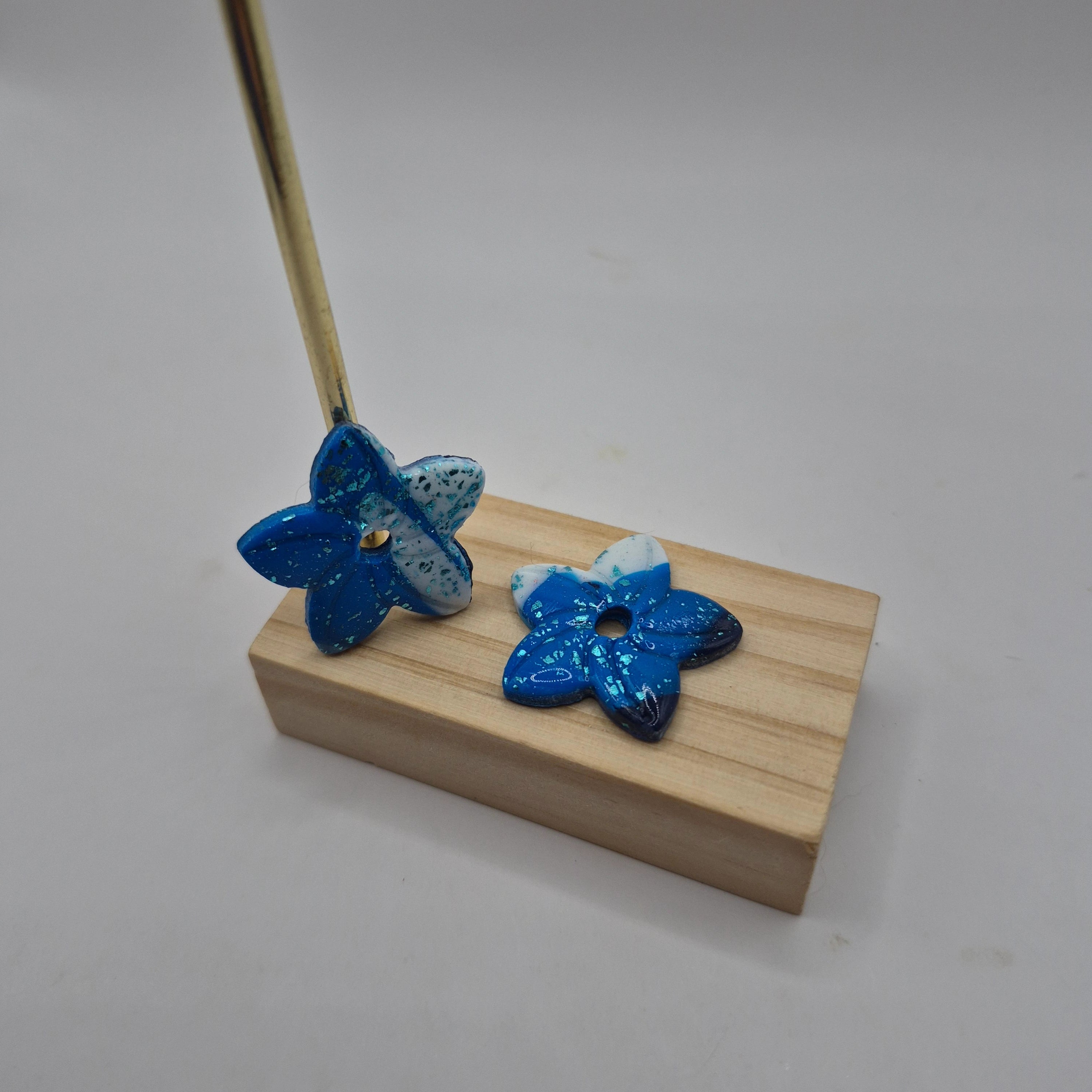Boucles d’oreilles pampilles en forme de fleur de tiaré, en pâte polymère bleu et blanc pailleté, faites main avec finition résine, présentées sur un support en bois — parure artisanale Thalia.