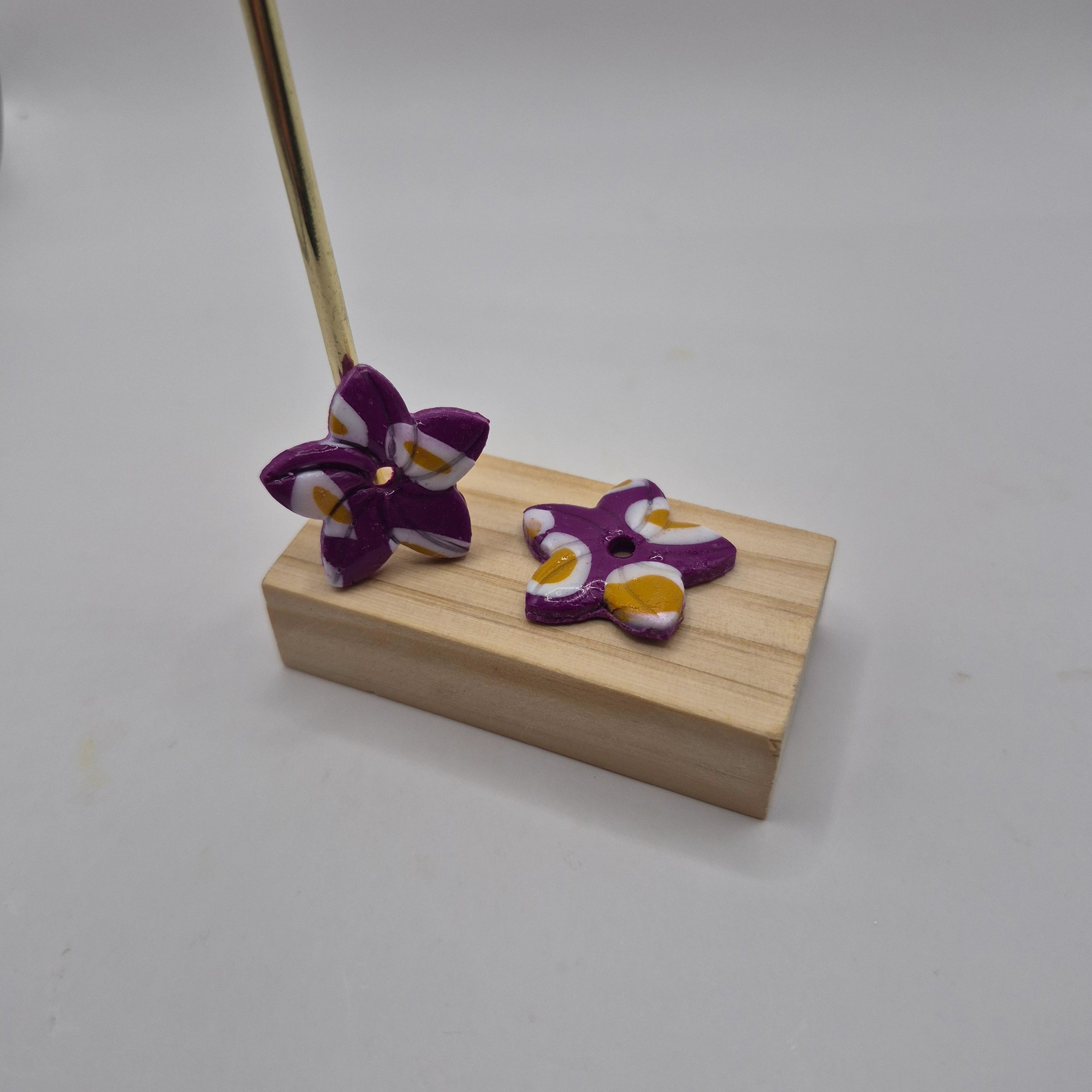 Boucles d’oreilles pampilles en forme de fleur de tiaré, en pâte polymère violette, blanche et ocre, faites main avec finition résine, présentées sur un support en bois — parure artisanale Thalia.