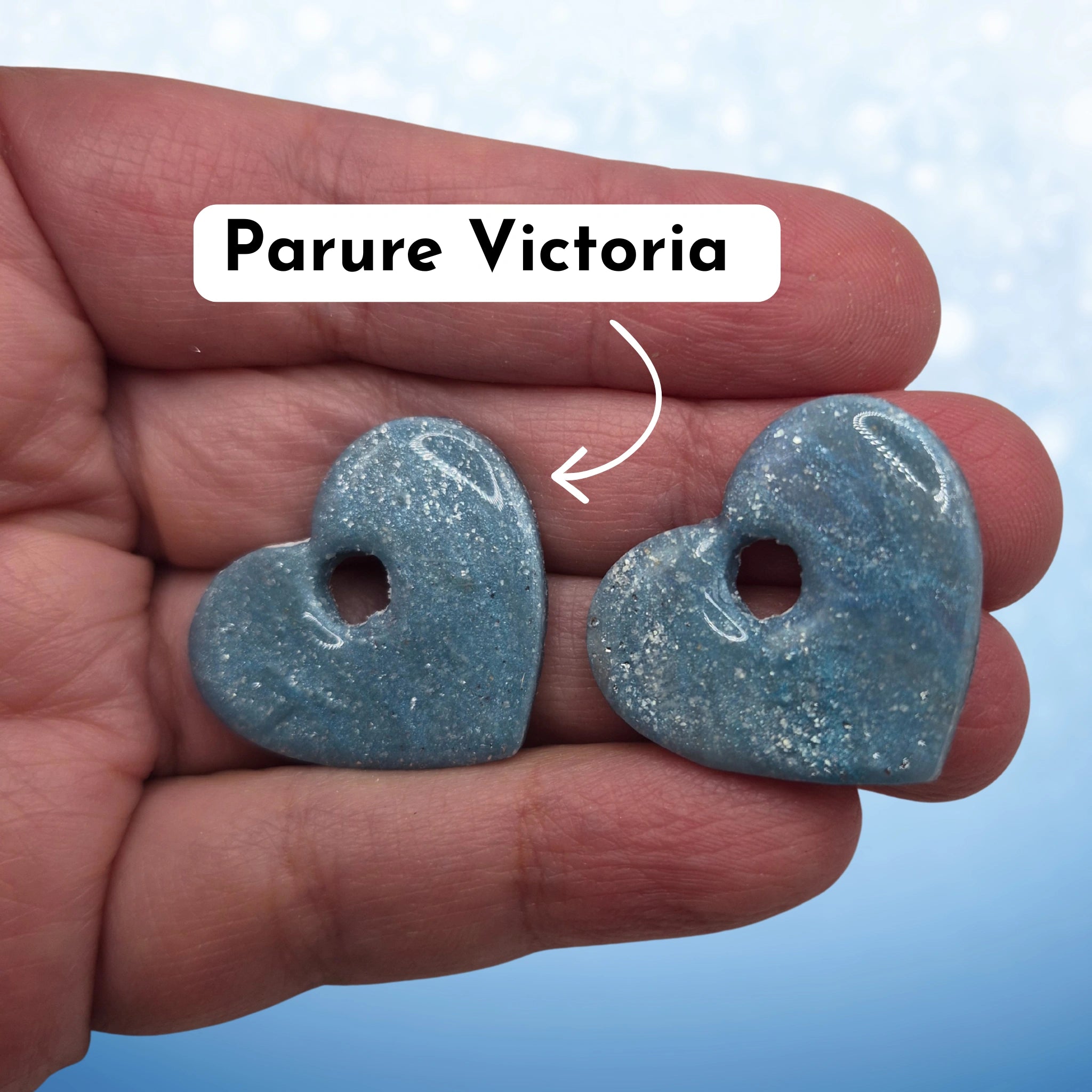Pampilles en pâte polymère bleues, en forme de cœur, avec l’inscription « Parure Victoria », présentées tenues dans une main, sur un fond en dégradé de bleu.