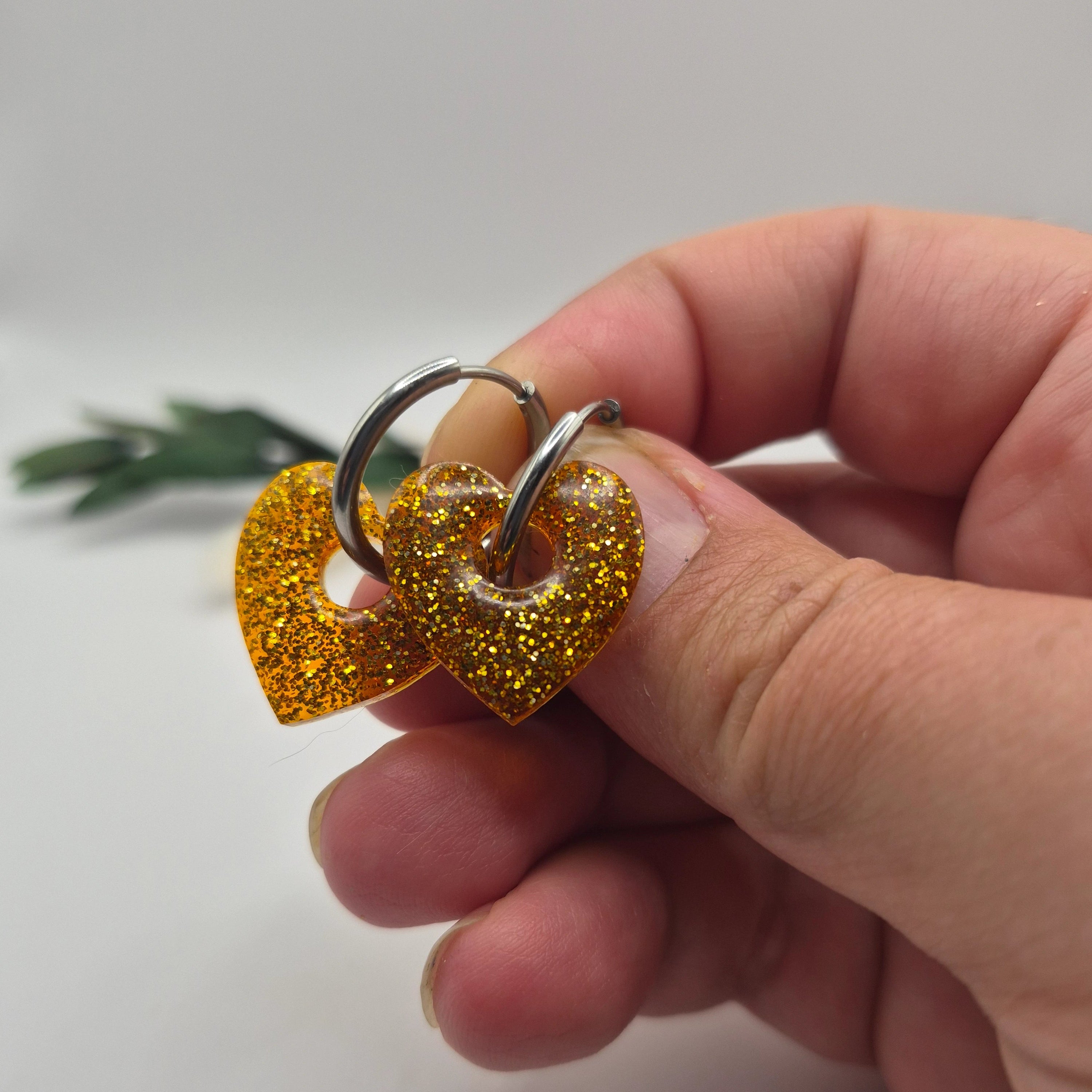 Pampilles Coralie dorées résine pailleté sur créoles inoxydables  argentées tenues à la main avec branche verte et pampa en fond de décor