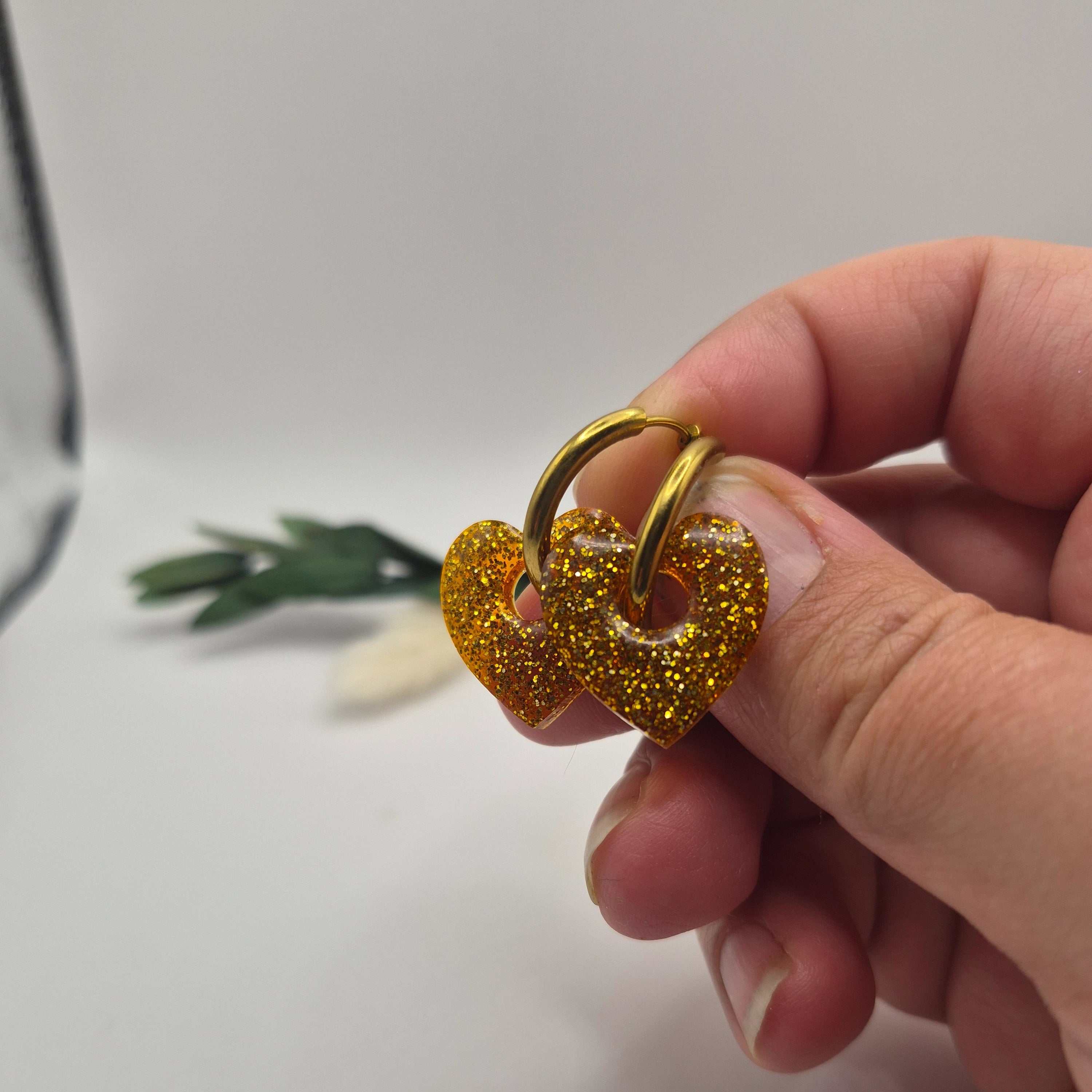 Pampilles Coralie dorées résine pailleté sur créoles inoxydables  dorées tenues à la main avec branche verte et pampa en fond de décor