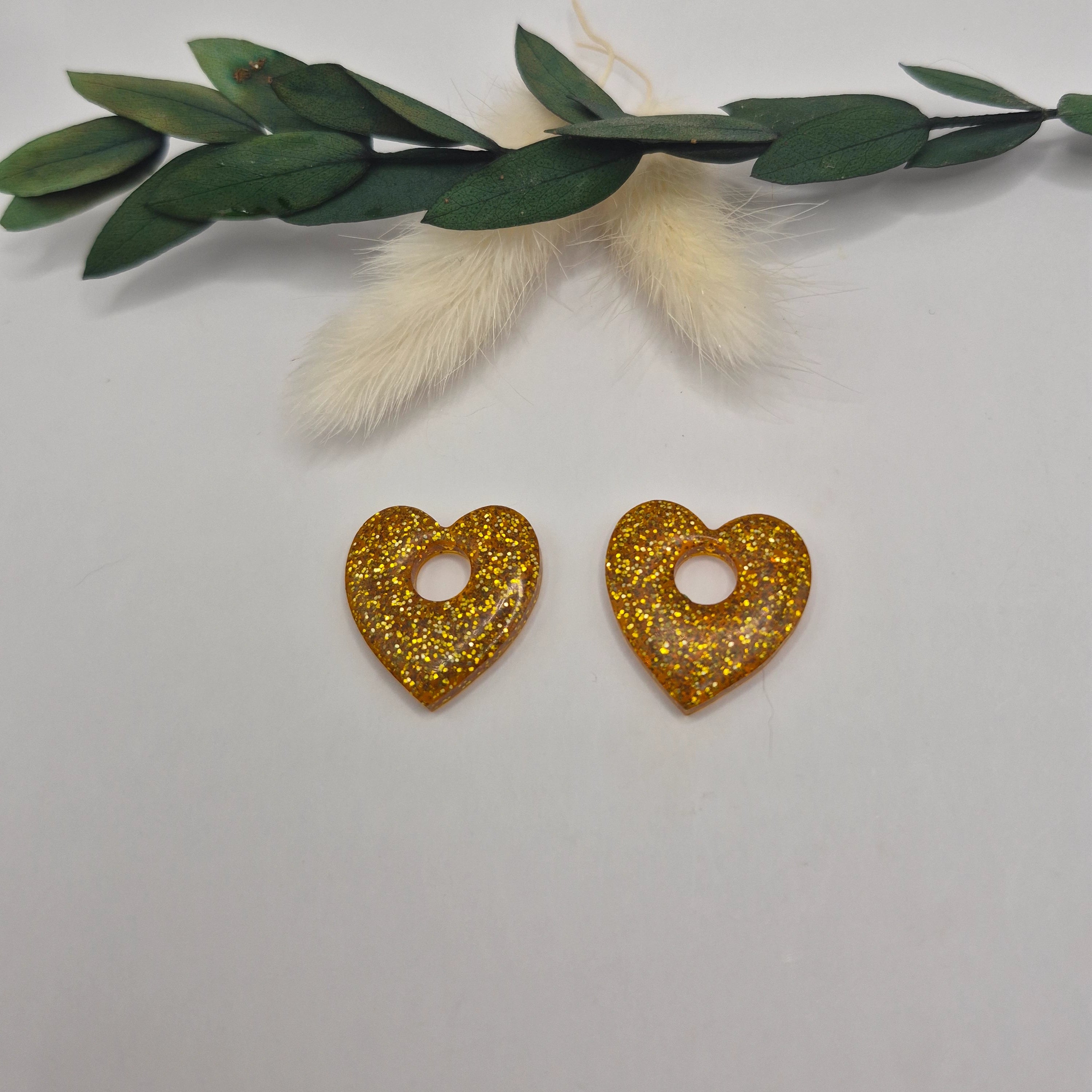 Pampilles Coralie dorées résine pailleté sur créoles inoxydables  posées à plat sur surface neutre avec décoration branche verte et pampa en fond de décor