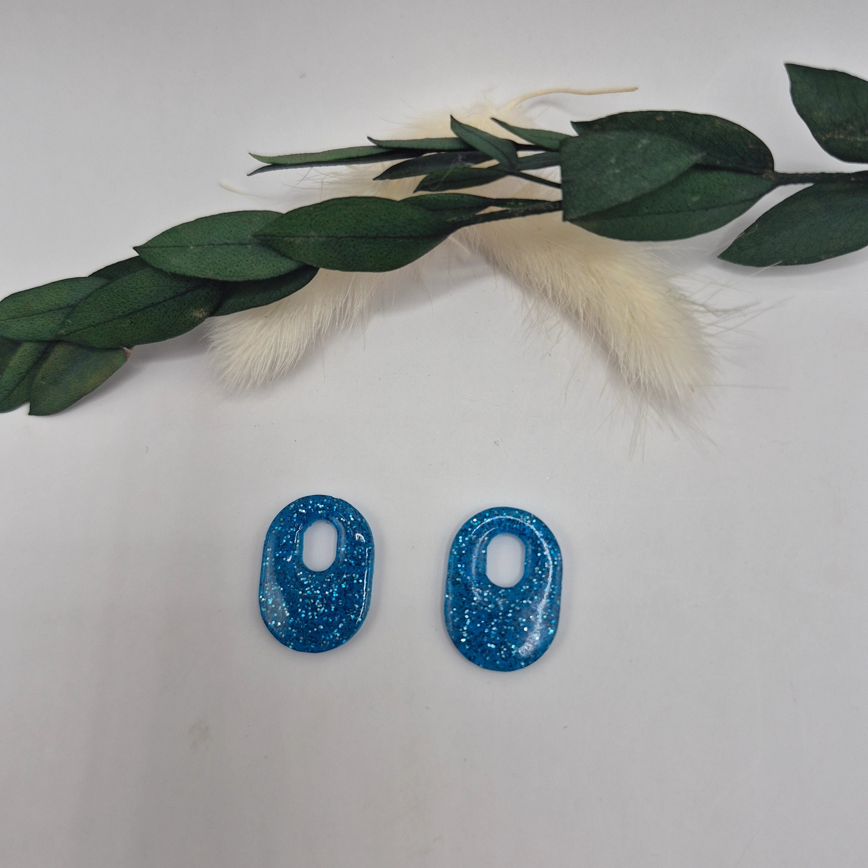 Boucles d’oreilles pampilles bleues Jessie en résine époxy pailletée posées à plat sur fond neutre avec branche de pampa et feuilles