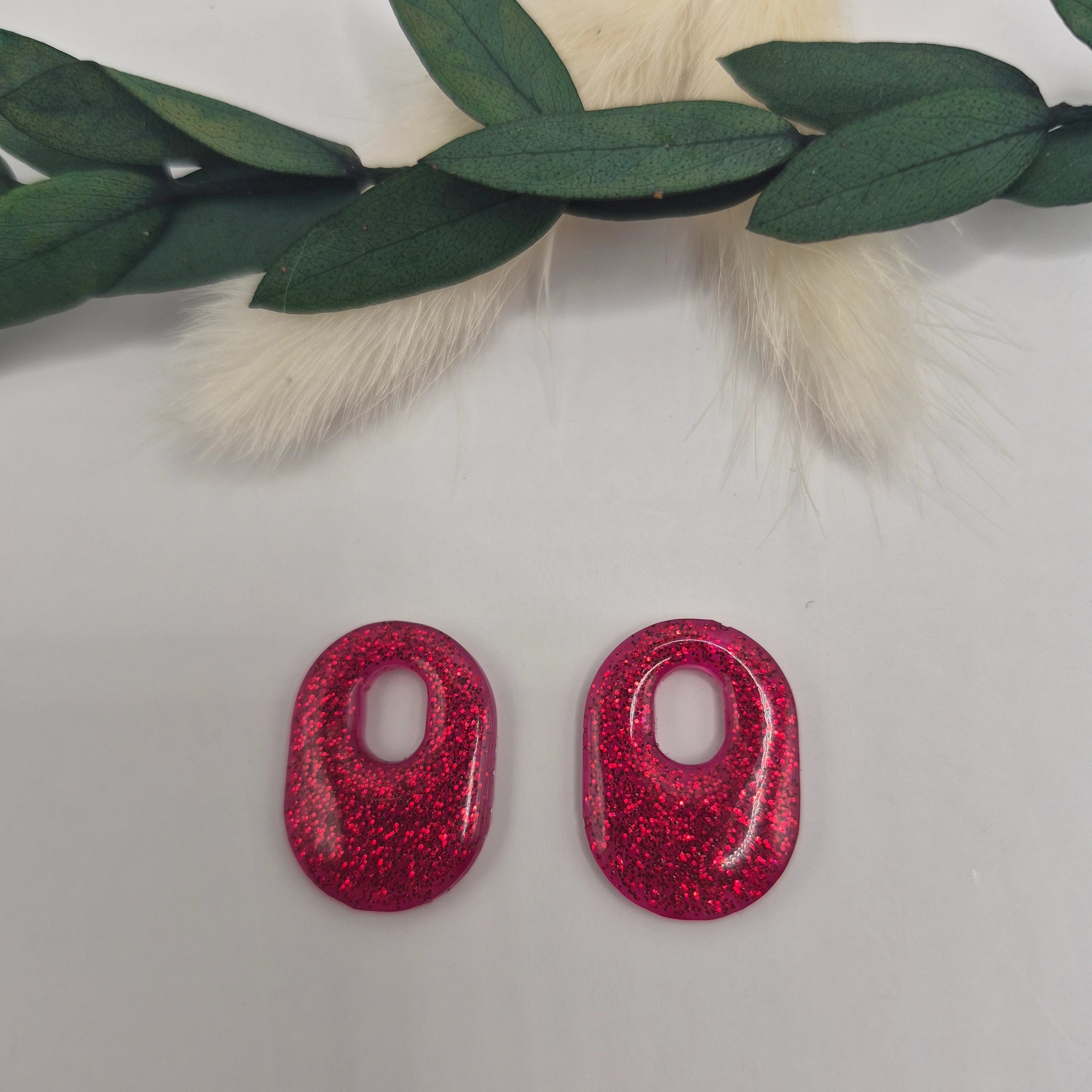 Boucles d’oreilles pampilles rouges Jessie en résine époxy pailletée posées à plat sur fond neutre avec branche de pampa et feuilles