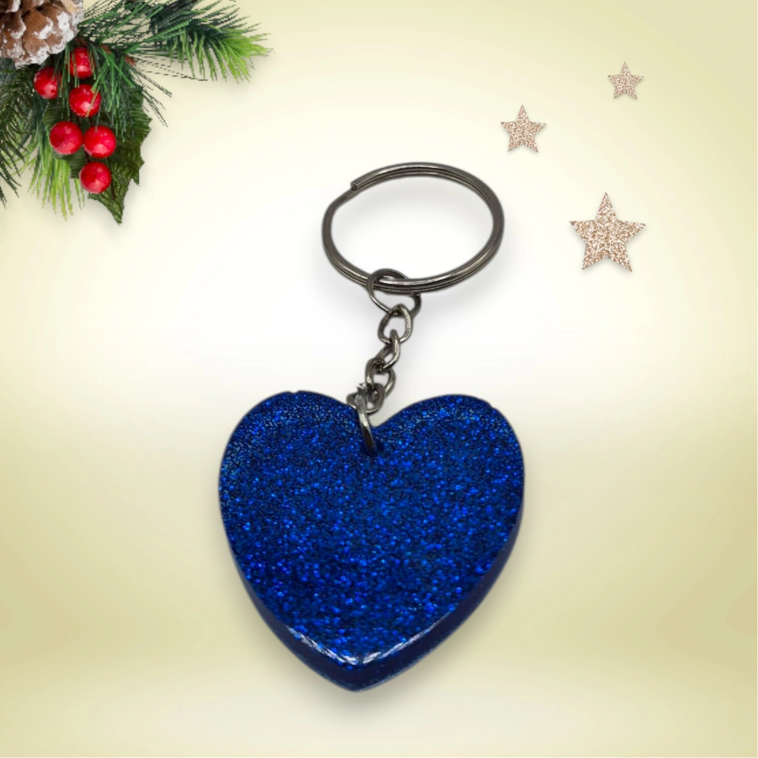 Porte-clés en résine bleue pailletée en forme de cœur, avec anneau argenté, posé sur un fond orné de branches de sapin et d’étoiles décoratives.