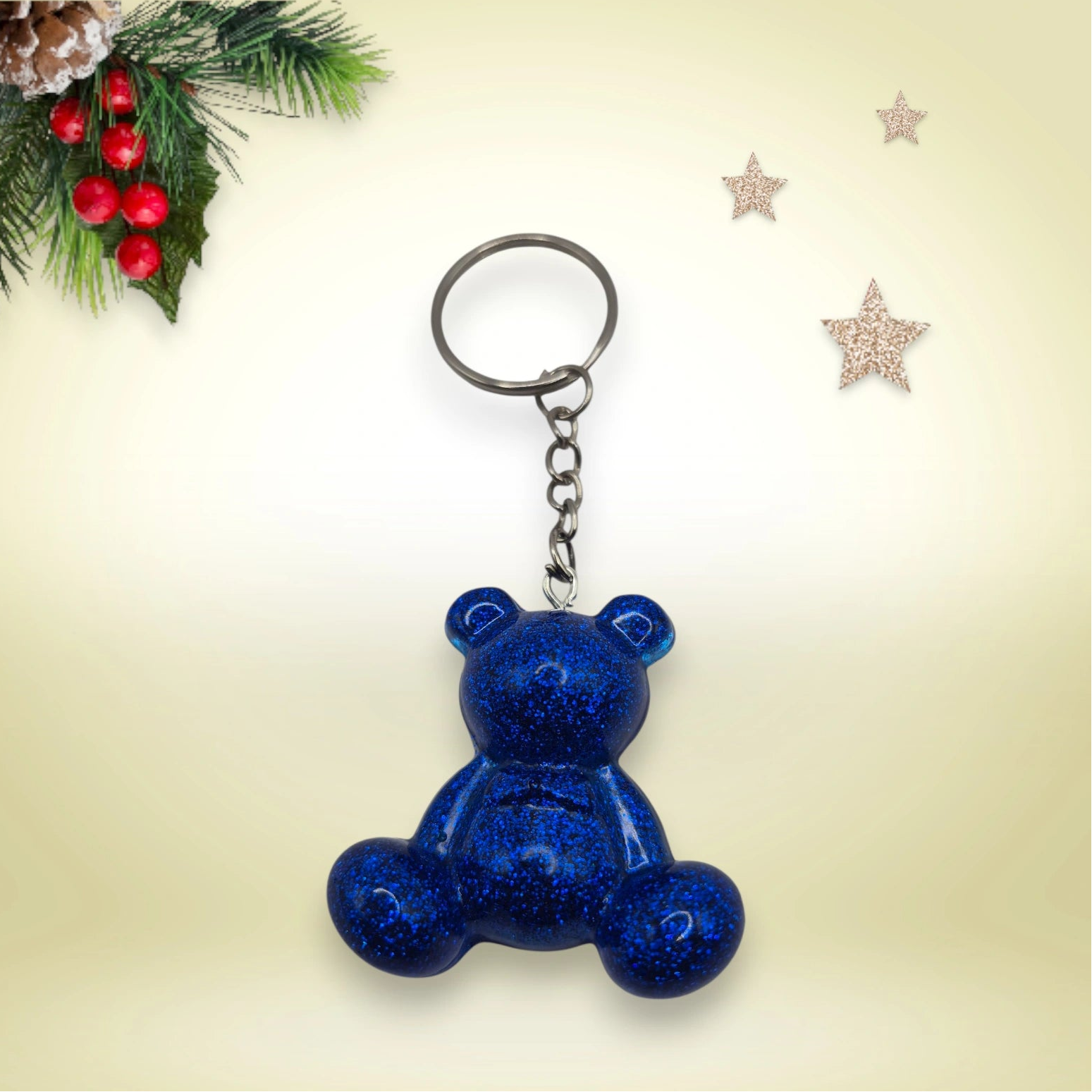 Porte-clés grand ourson en résine bleue foncé pailletée, avec anneau argenté, posé sur un fond orné de branches de sapin et d’étoiles décoratives.