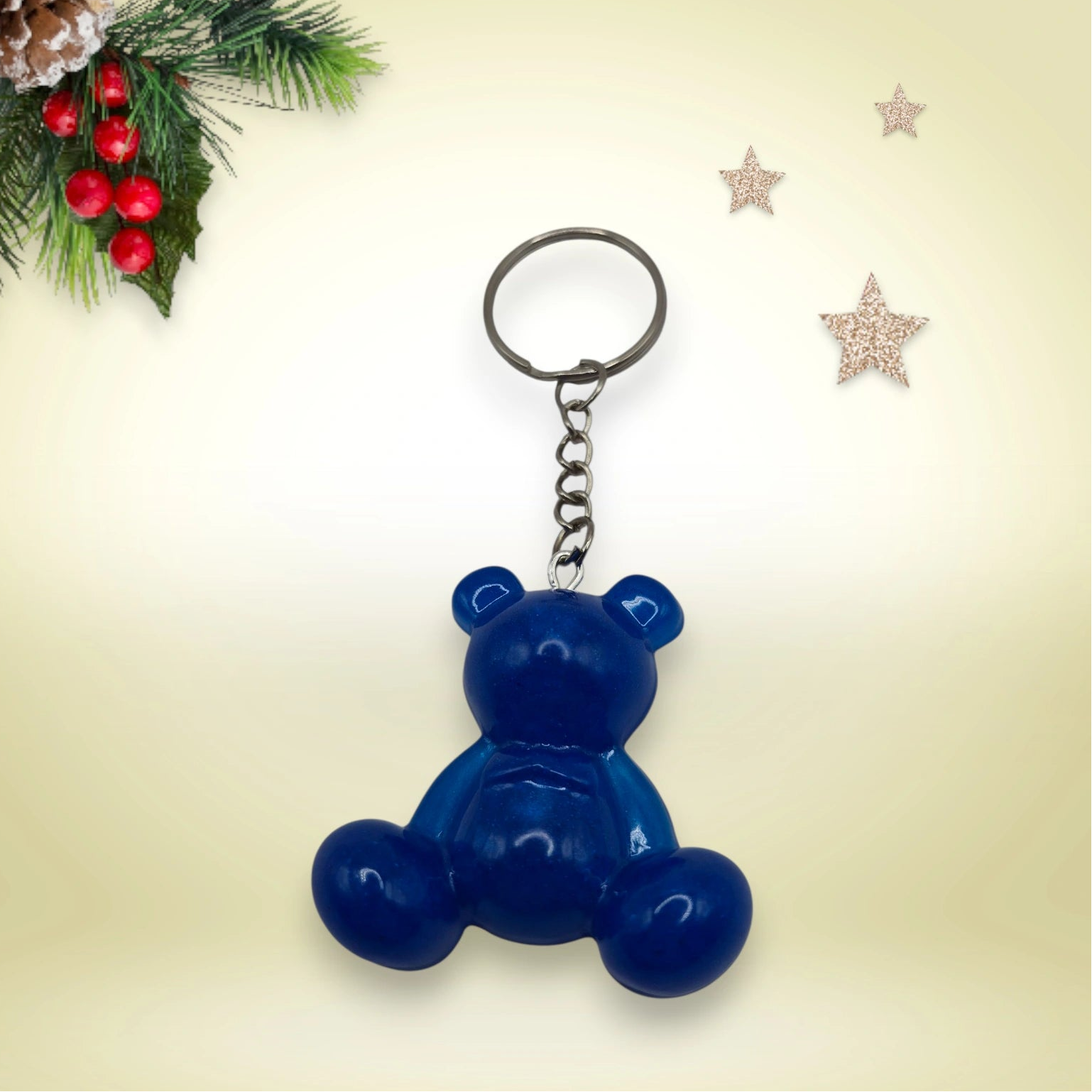 Porte-clés grand ourson en résine bleue, avec anneau argenté, posé sur un fond orné de branches de sapin et d’étoiles décoratives.