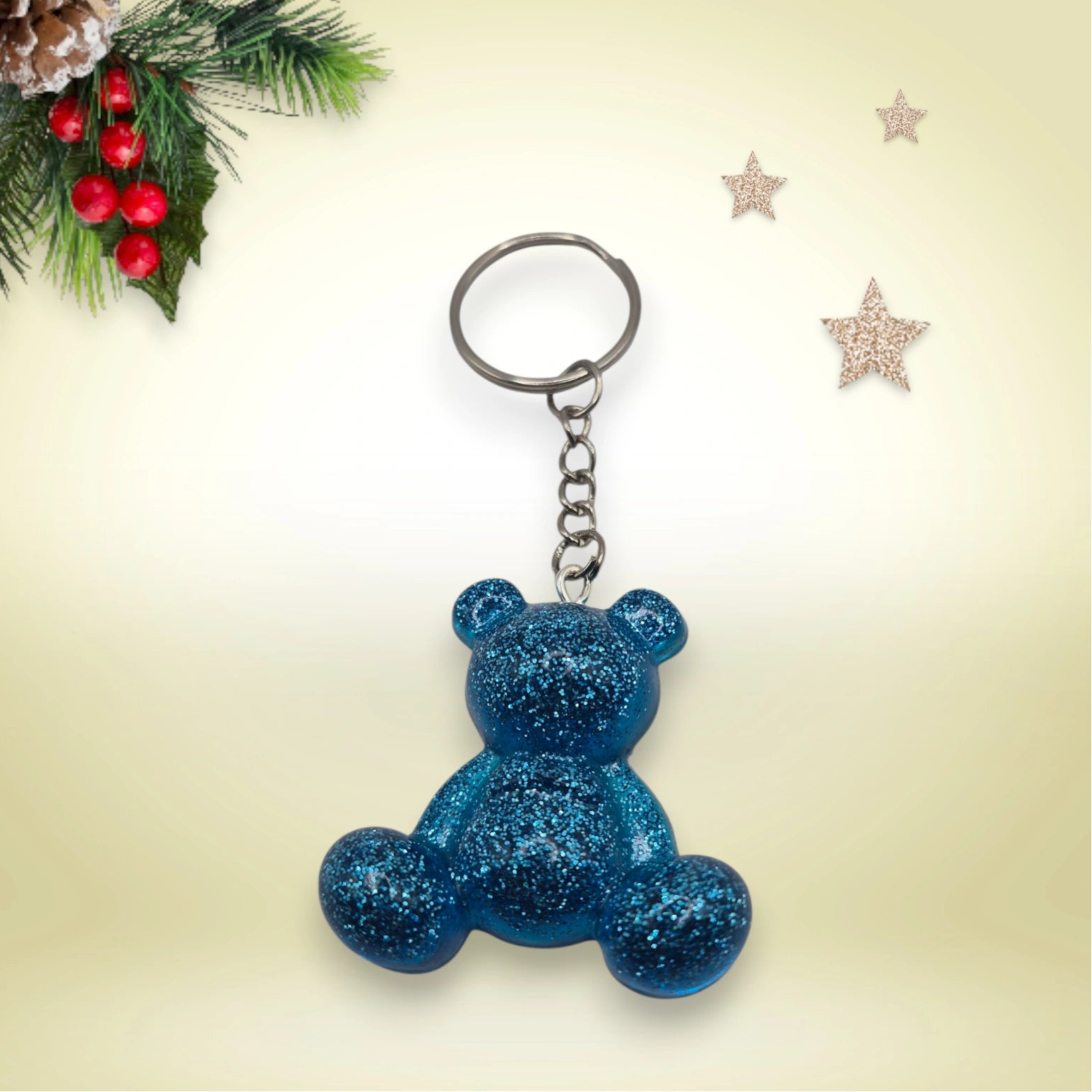 Porte-clés grand ourson en résine bleue pailletée nacré, avec anneau argenté, posé sur un fond orné de branches de sapin et d’étoiles décoratives.