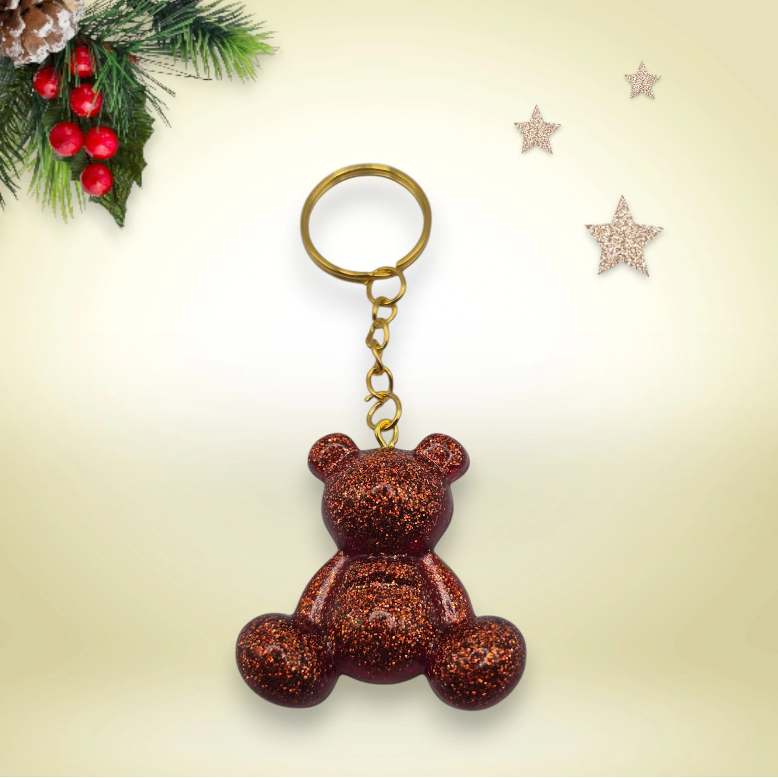 Porte-clés grand ourson en résine cuivre pailletée, avec anneau doré, posé sur un fond orné de branches de sapin et d’étoiles décoratives.