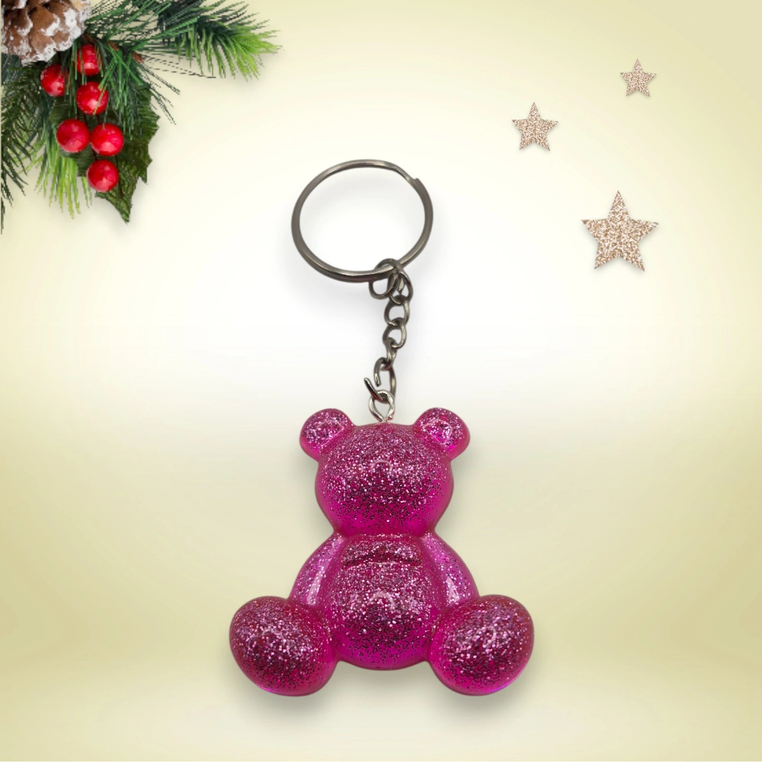 Porte-clés grand ourson en résine rose pailletée, avec anneau argenté, posé sur un fond orné de branches de sapin et d’étoiles décoratives.