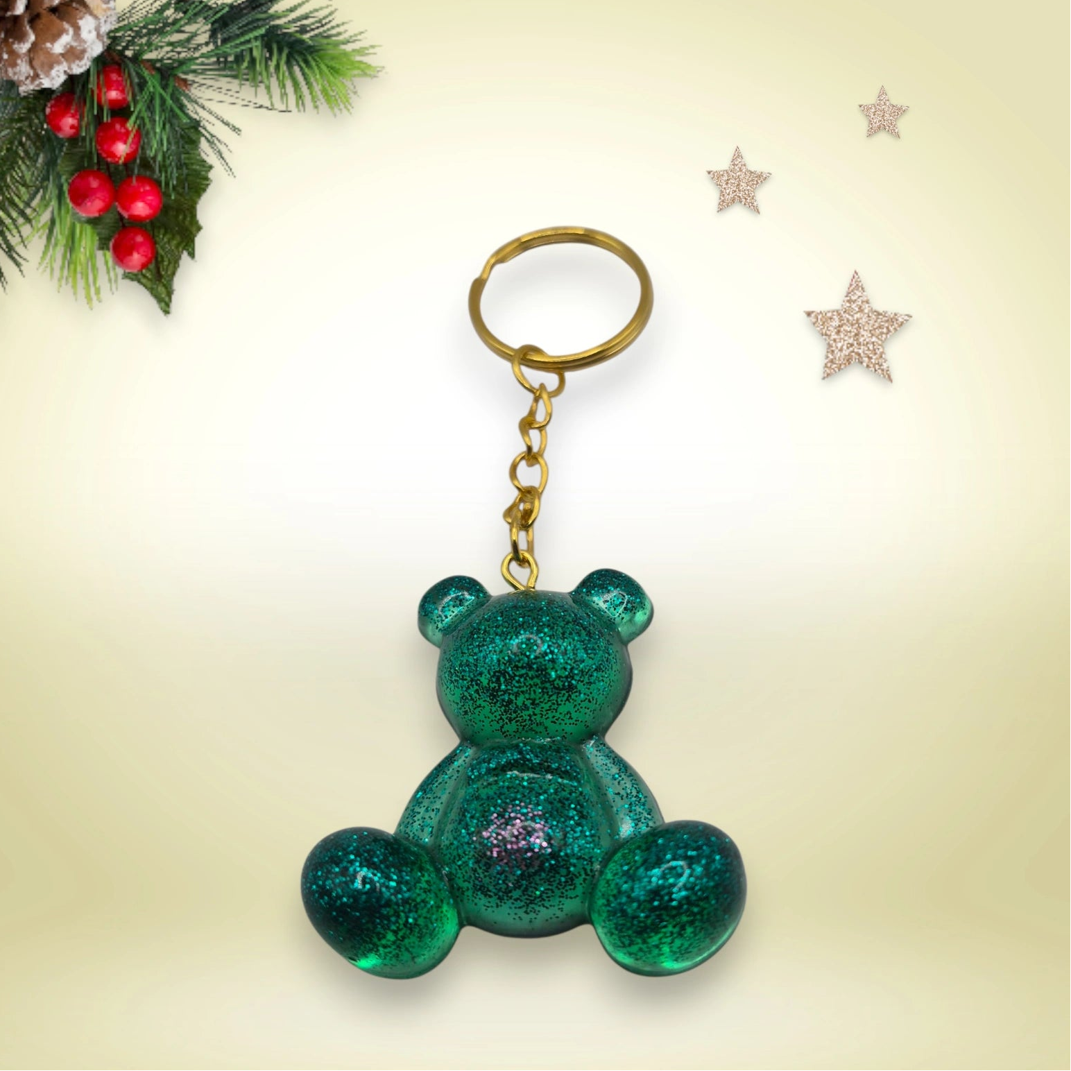 Porte-clés grand ourson en résine verte pailletée, avec anneau doré, posé sur un fond orné de branches de sapin et d’étoiles décoratives.