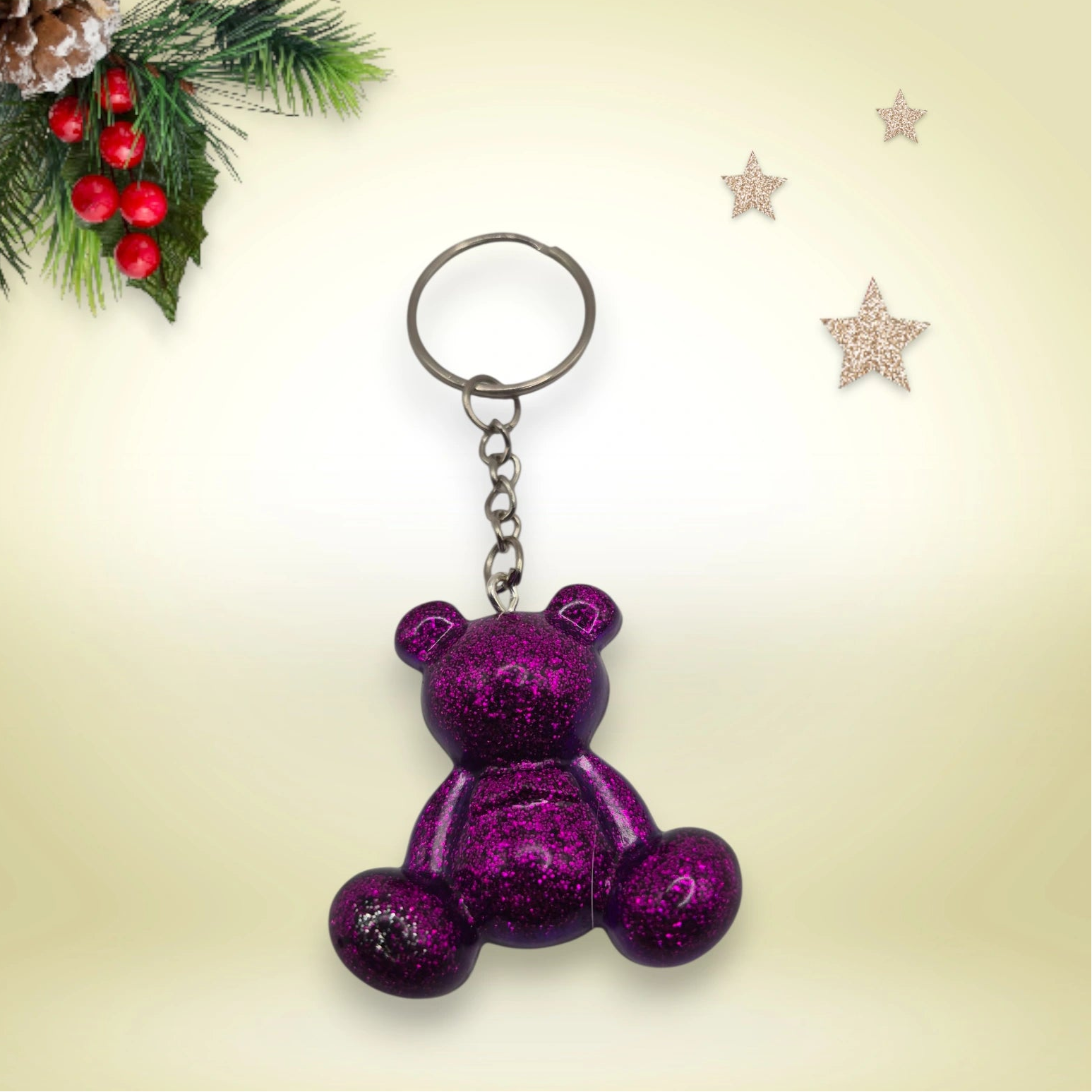 Porte-clés grand ourson en résine violette pailletée, avec anneau argenté, posé sur un fond orné de branches de sapin et d’étoiles décoratives.