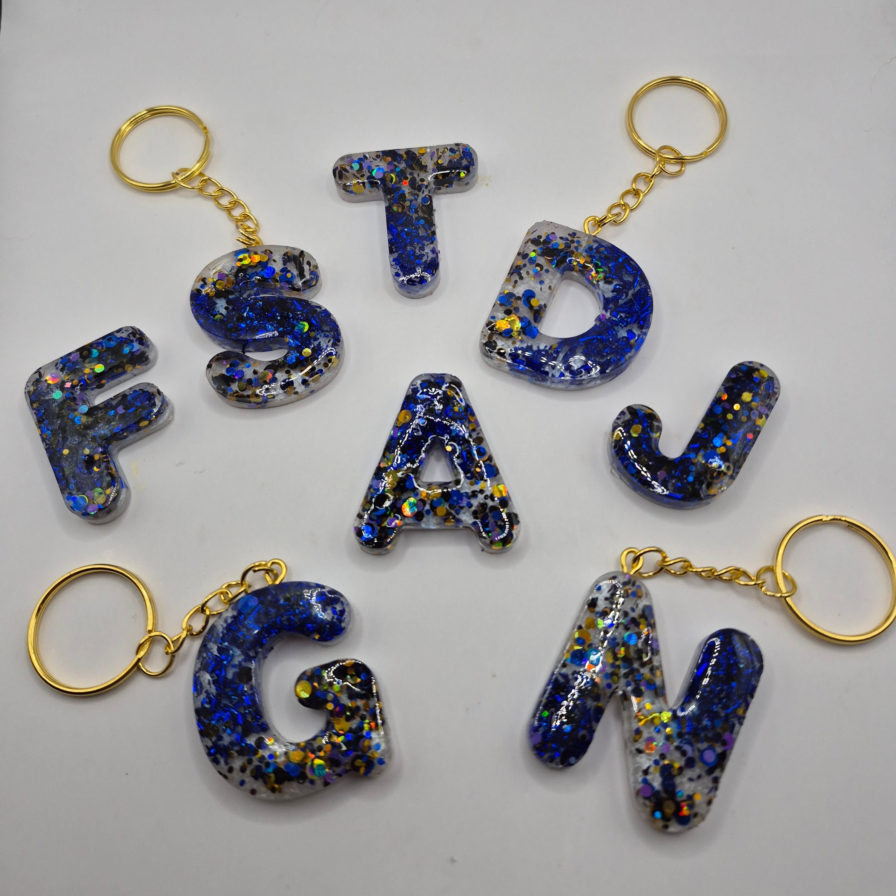 Porte-clés en forme de lettres de l’alphabet, en résine bleue pailletée, présentés à plat avec leurs anneaux dorés.