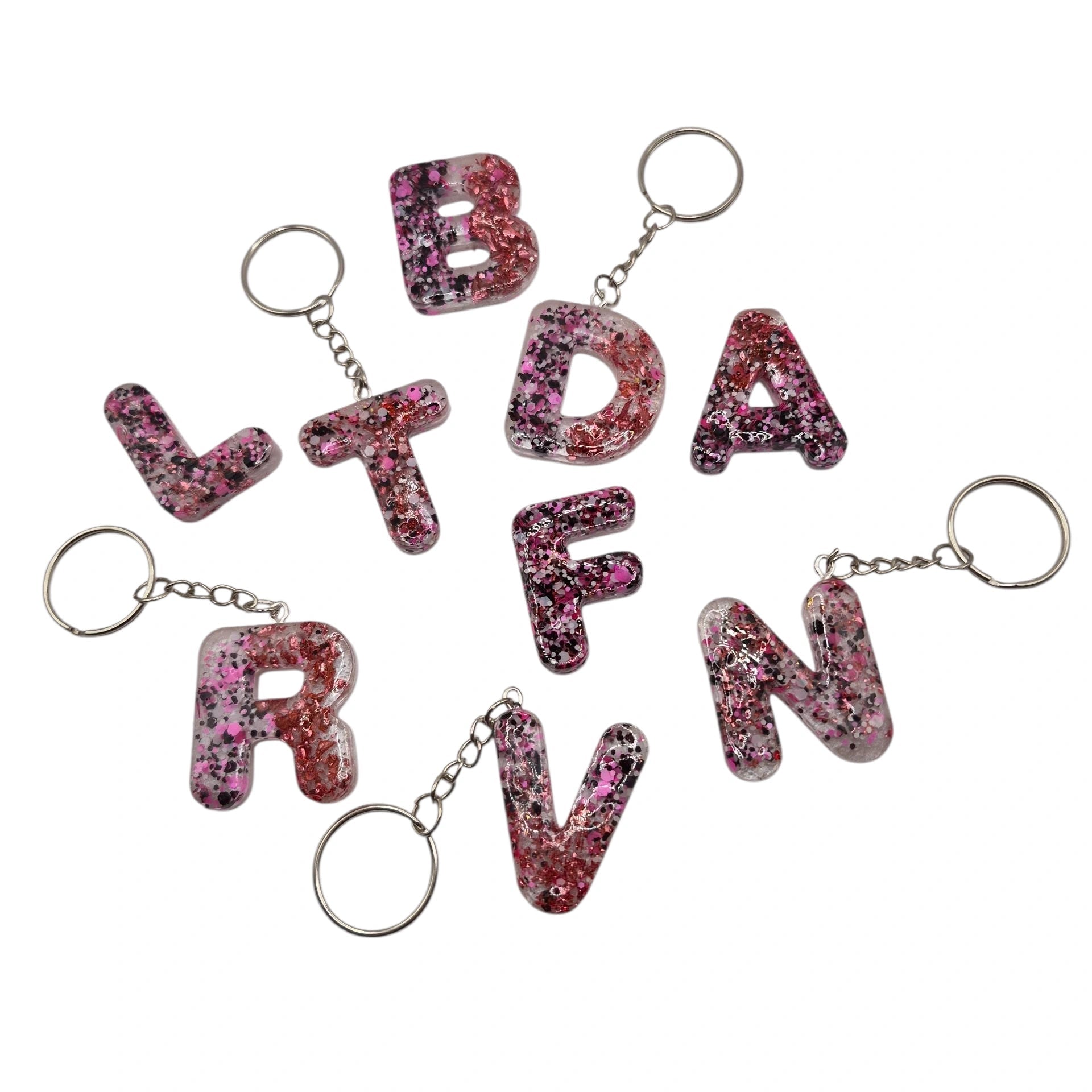 Porte-clés en forme de lettres de l’alphabet, en résine rose pailletée, présentés à plat avec leurs anneaux argentés.