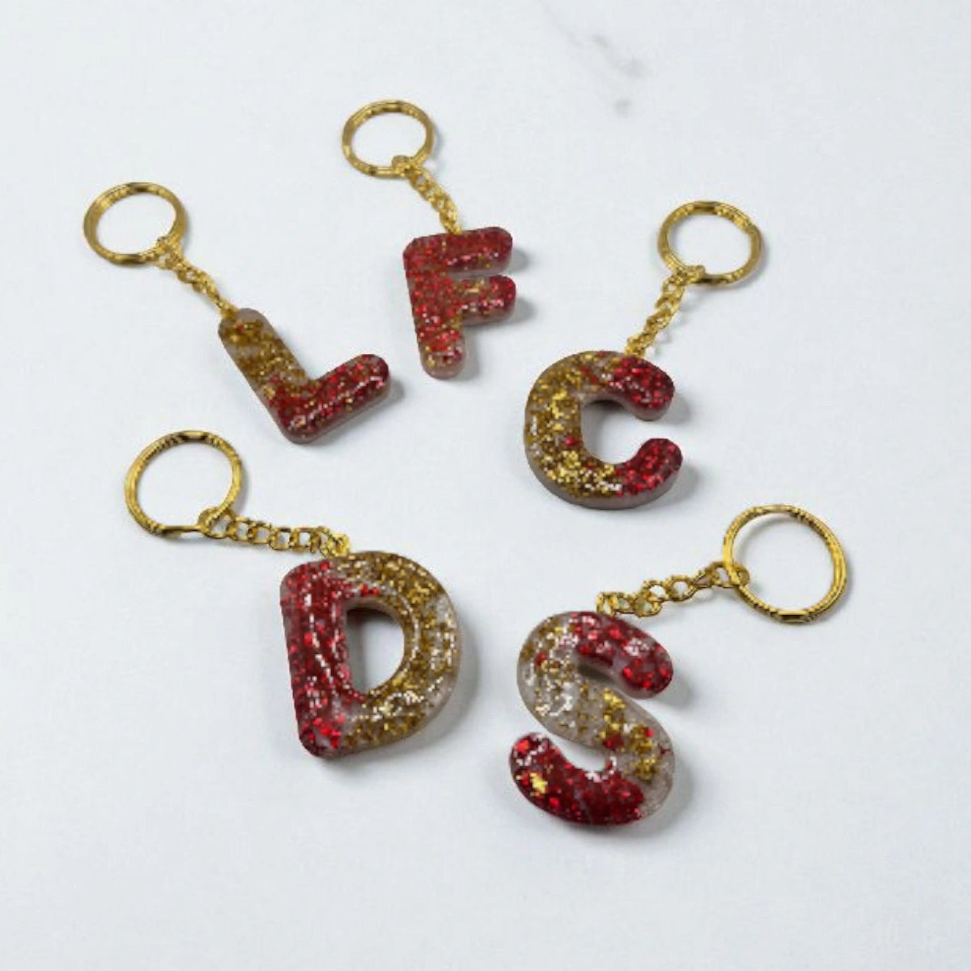 Porte-clés en forme de lettres de l’alphabet, en résine rouge et doré pailletée, présentés à plat avec leurs anneaux dorés.