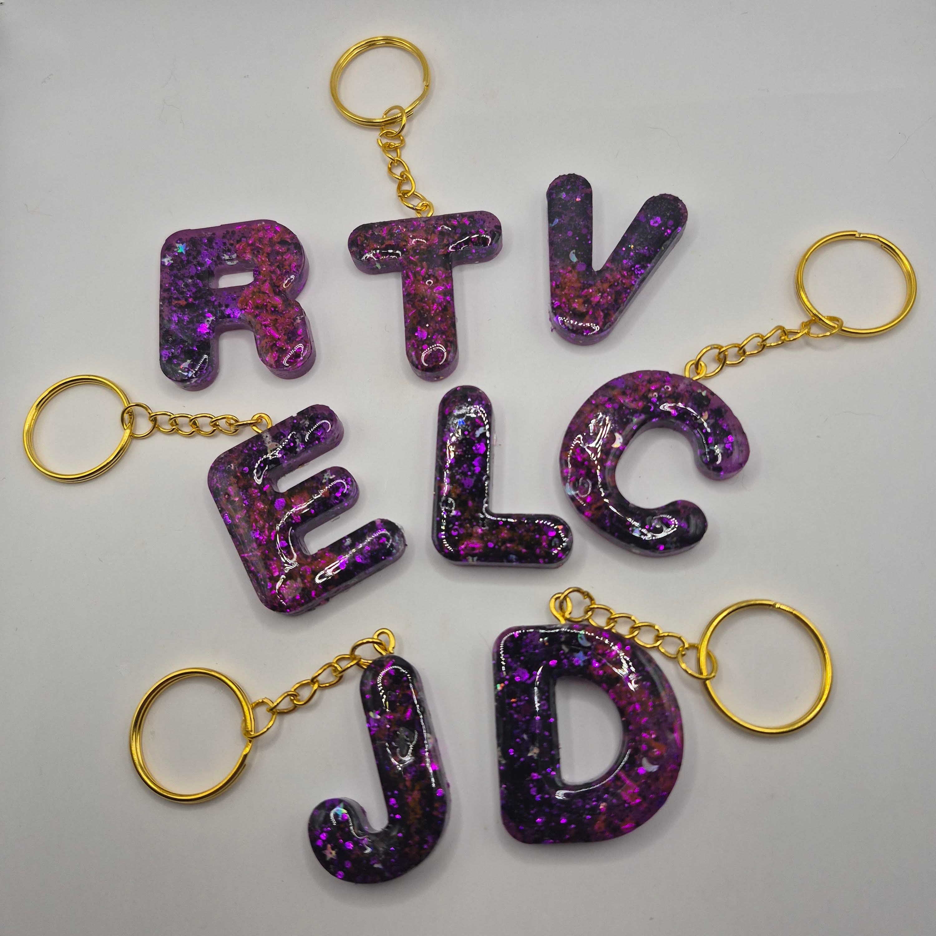 Porte-clés en forme de lettres de l’alphabet, en résine violet, rose pailletée, présentés à plat avec leurs anneaux dorés.
