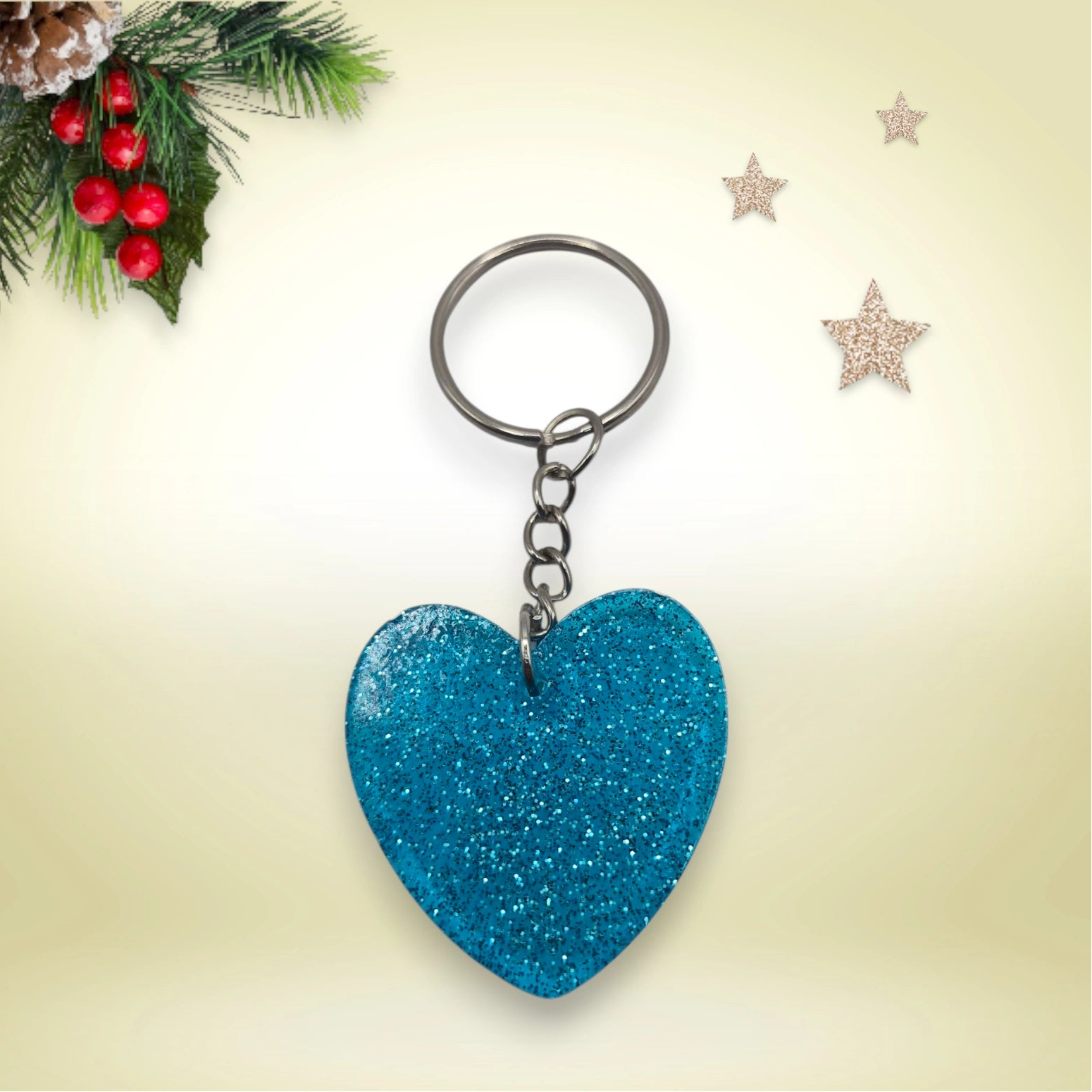 Porte-clés en résine bleue pailletée en forme de cœur, avec anneau argenté, posé sur un fond orné de branches de sapin et d’étoiles décoratives.