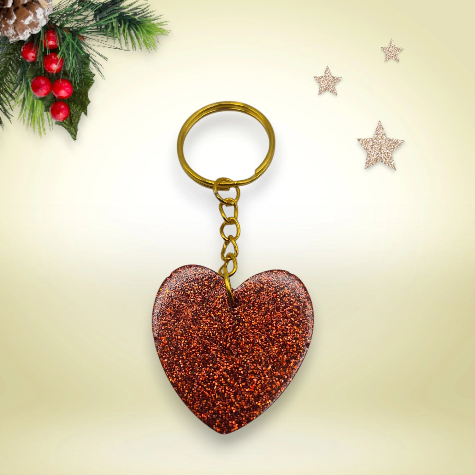 Porte-clés en résine rouge pailletée en forme de cœur, avec anneau doré, posé sur un fond orné de branches de sapin et d’étoiles décoratives.