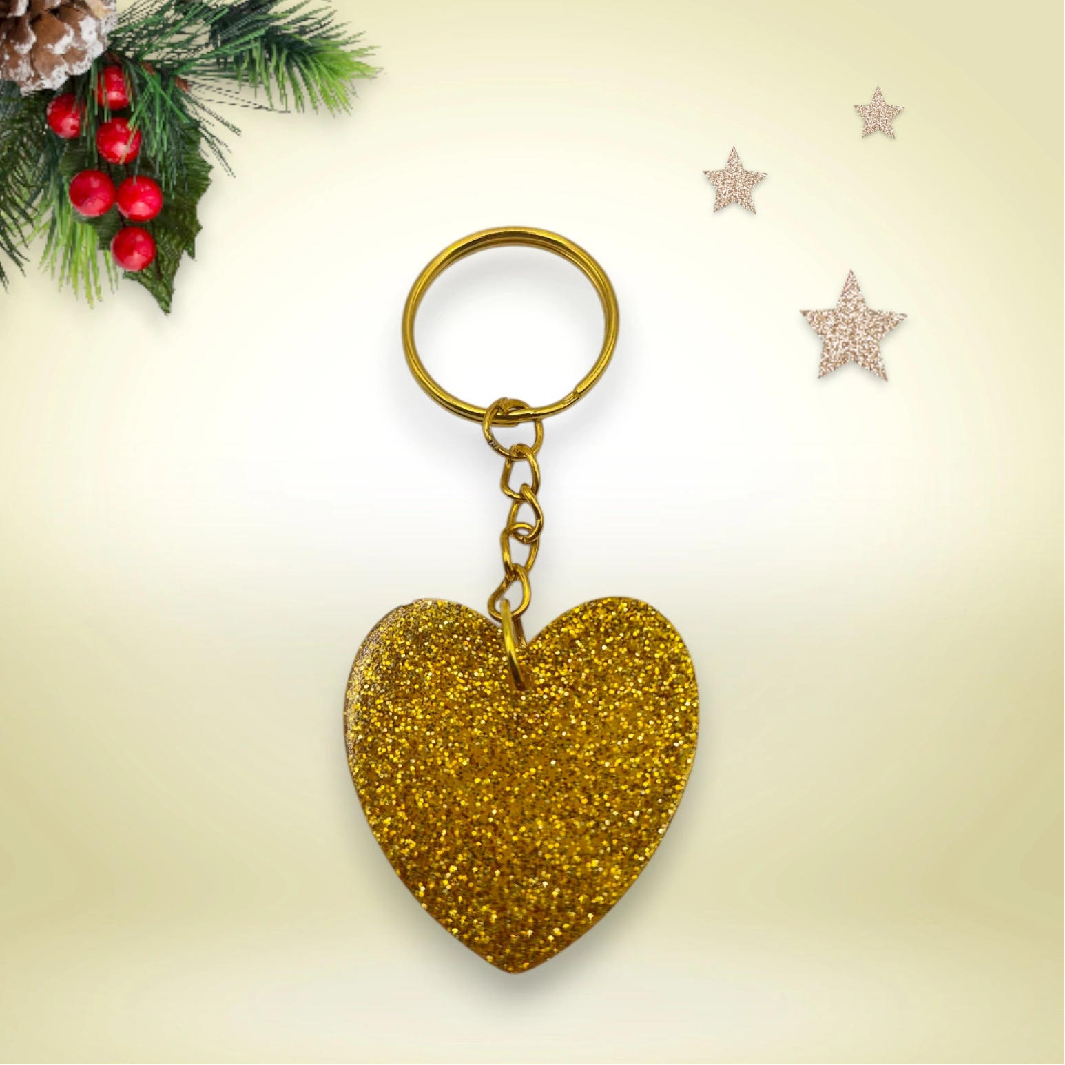 Porte-clés en résine doré pailletée en forme de cœur, avec anneau doré, posé sur un fond orné de branches de sapin et d’étoiles décoratives.
