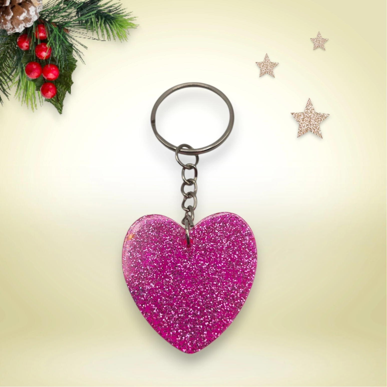Porte-clés en résine rose pailletée en forme de cœur, avec anneau argenté, posé sur un fond orné de branches de sapin et d’étoiles décoratives.