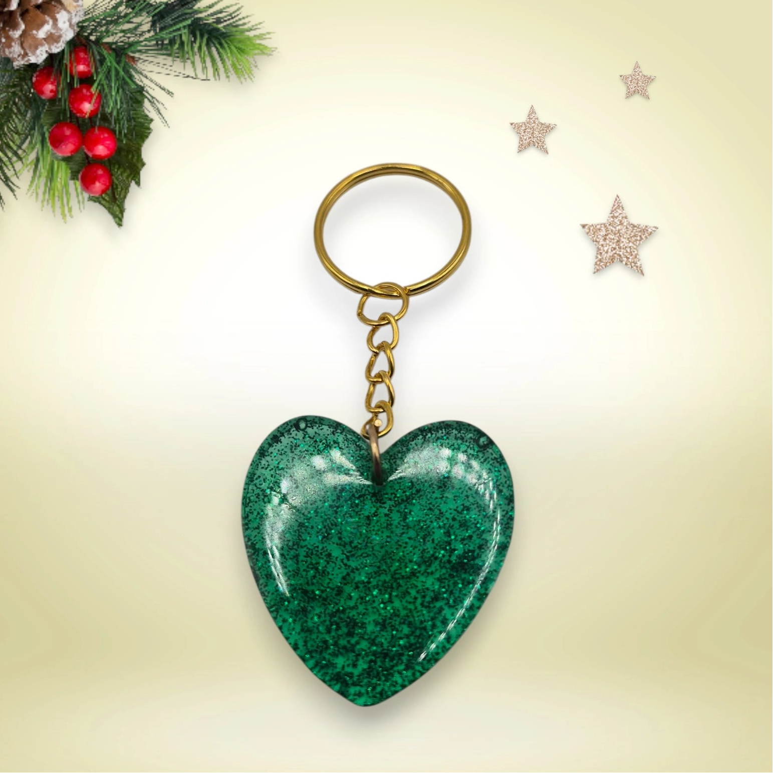 Porte-clés en résine verte pailletée en forme de cœur, avec anneau doré, posé sur un fond orné de branches de sapin et d’étoiles décoratives.