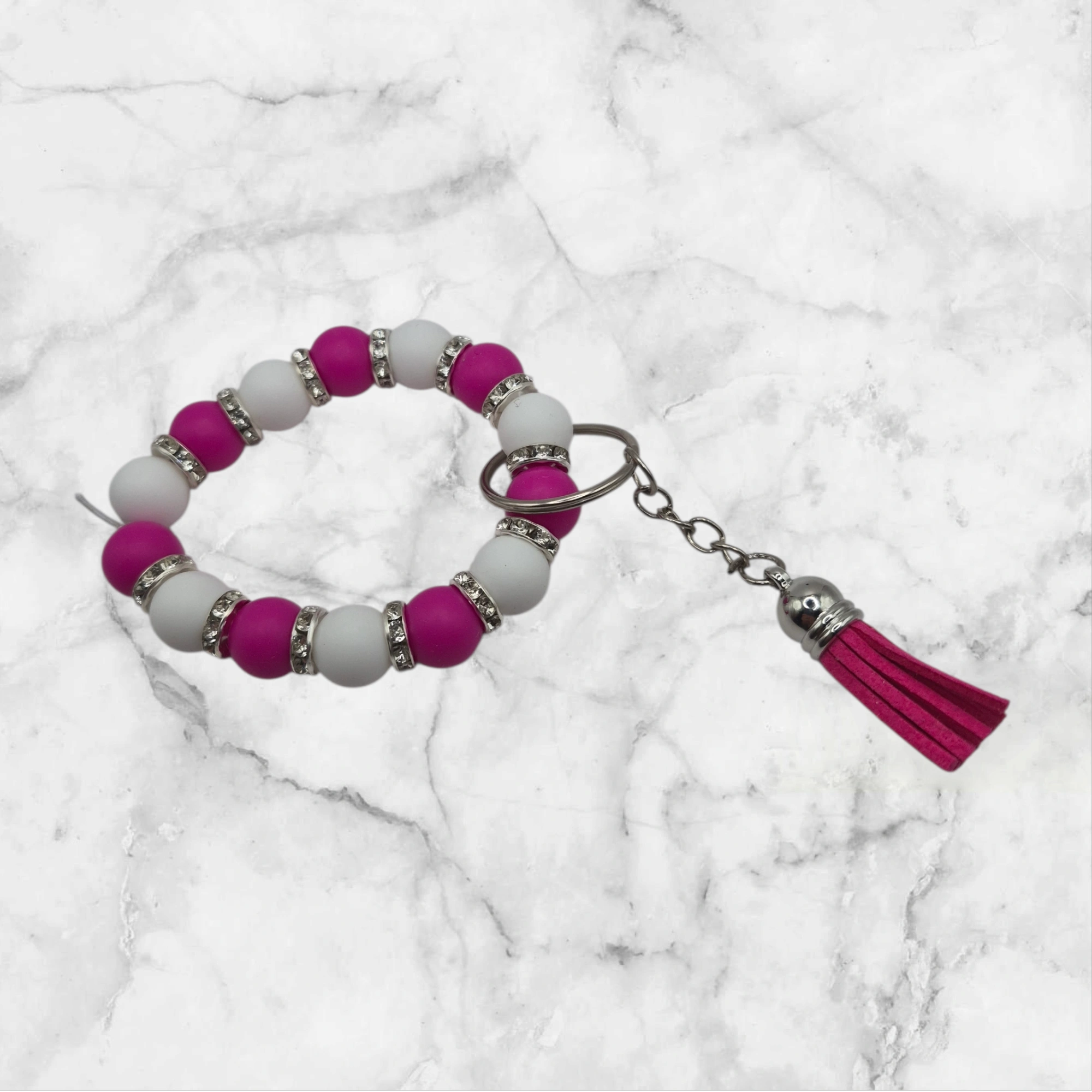 Porte-clé anneau en perles de silicone (7 cm de diamètre) de couleur rose et blanc nacré, séparées par des perles d'espacement métalliques avec strass. L'accessoire est complété par un petit pompon fuchsia de 3 cm et un anneau argent.
