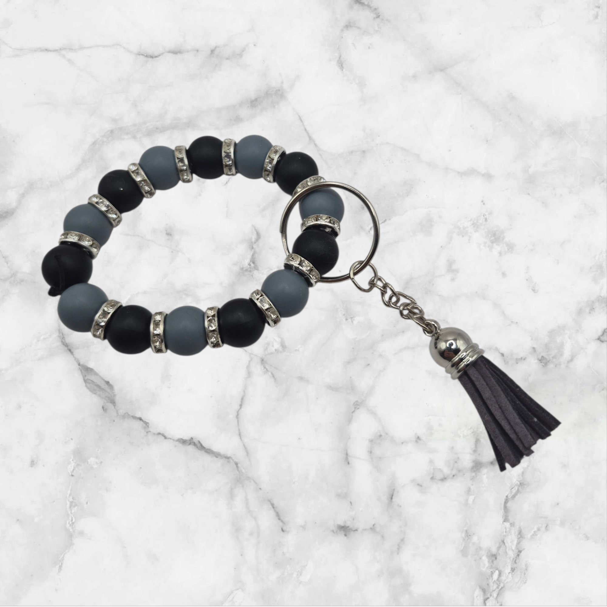 Porte-clé anneau en perles de silicone (7 cm de diamètre) de couleur noir et gris, séparées par des perles d'espacement métalliques avec strass. L'accessoire est complété par un petit pompon gris de 3 cm et un anneau argent.