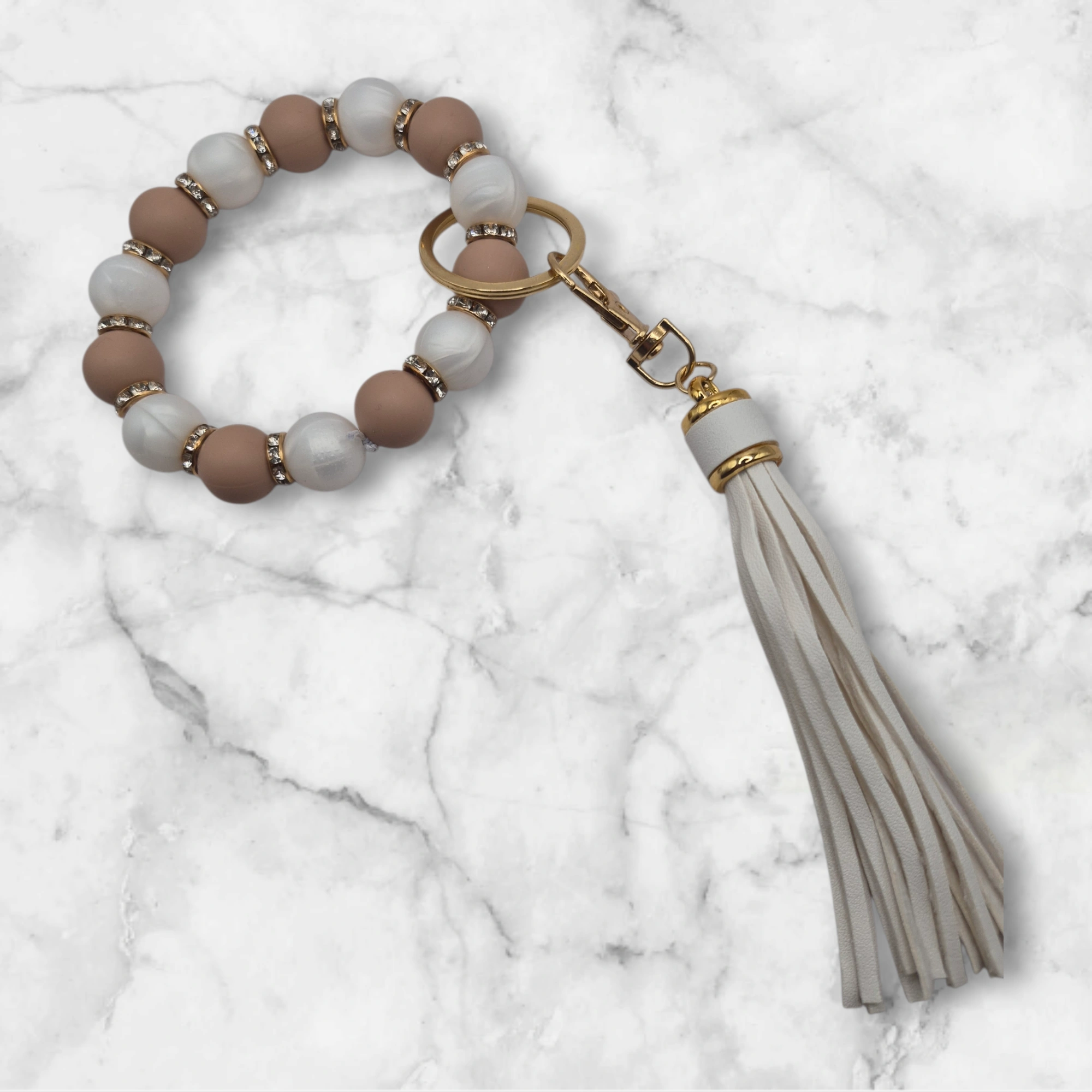 Porte-clé en perles de silicone blanc nacré et beige agrémenté de perles d'espacement métalliques incrustées de strass. Il est doté d'un anneau brisé et d'un mousqueton dorés, ainsi que d'un pompon décoratif blanc