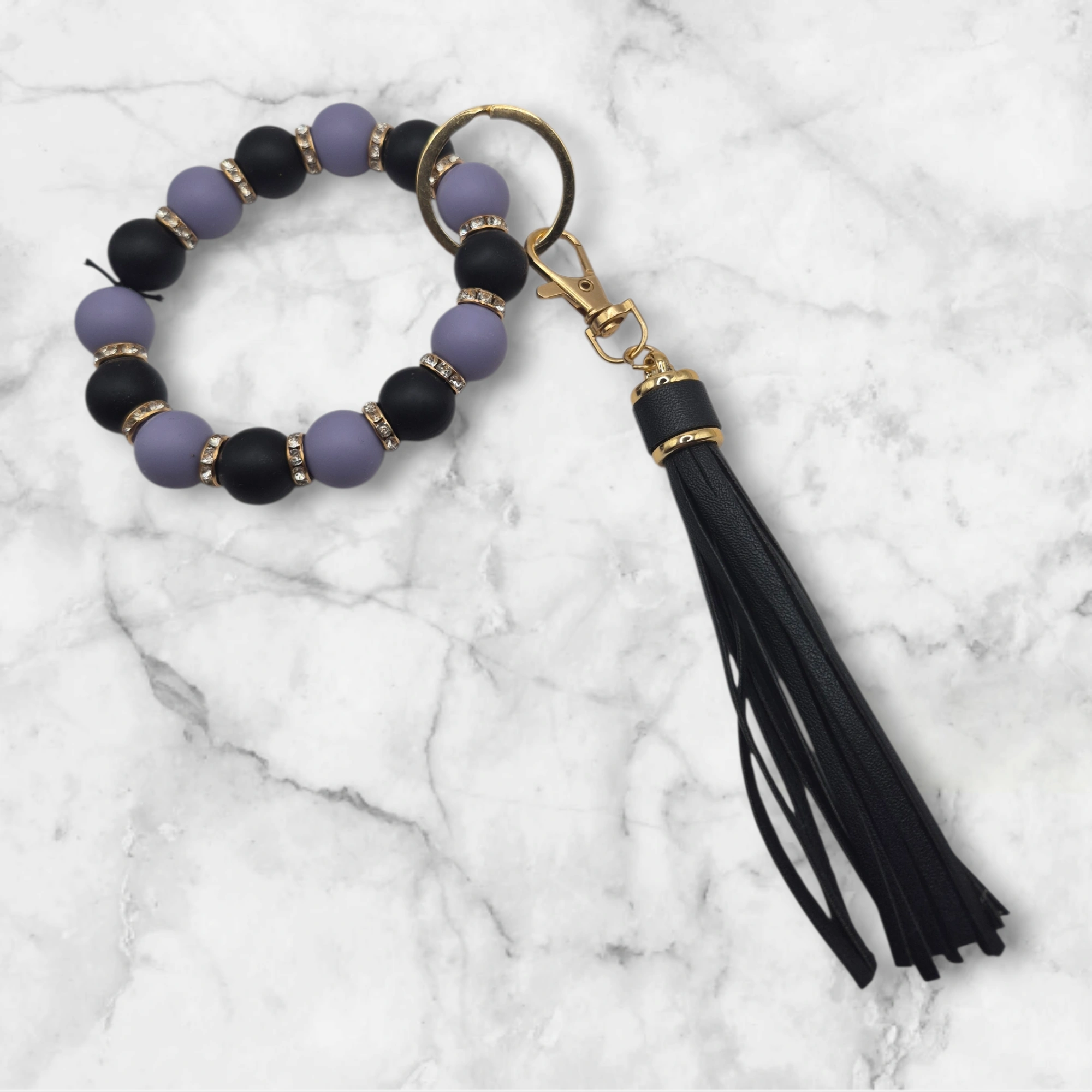 Porte-clé en perles de silicone noir et violet agrémenté de perles d'espacement métalliques incrustées de strass. Il est doté d'un anneau brisé et d'un mousqueton dorés, ainsi que d'un pompon décoratif noir.