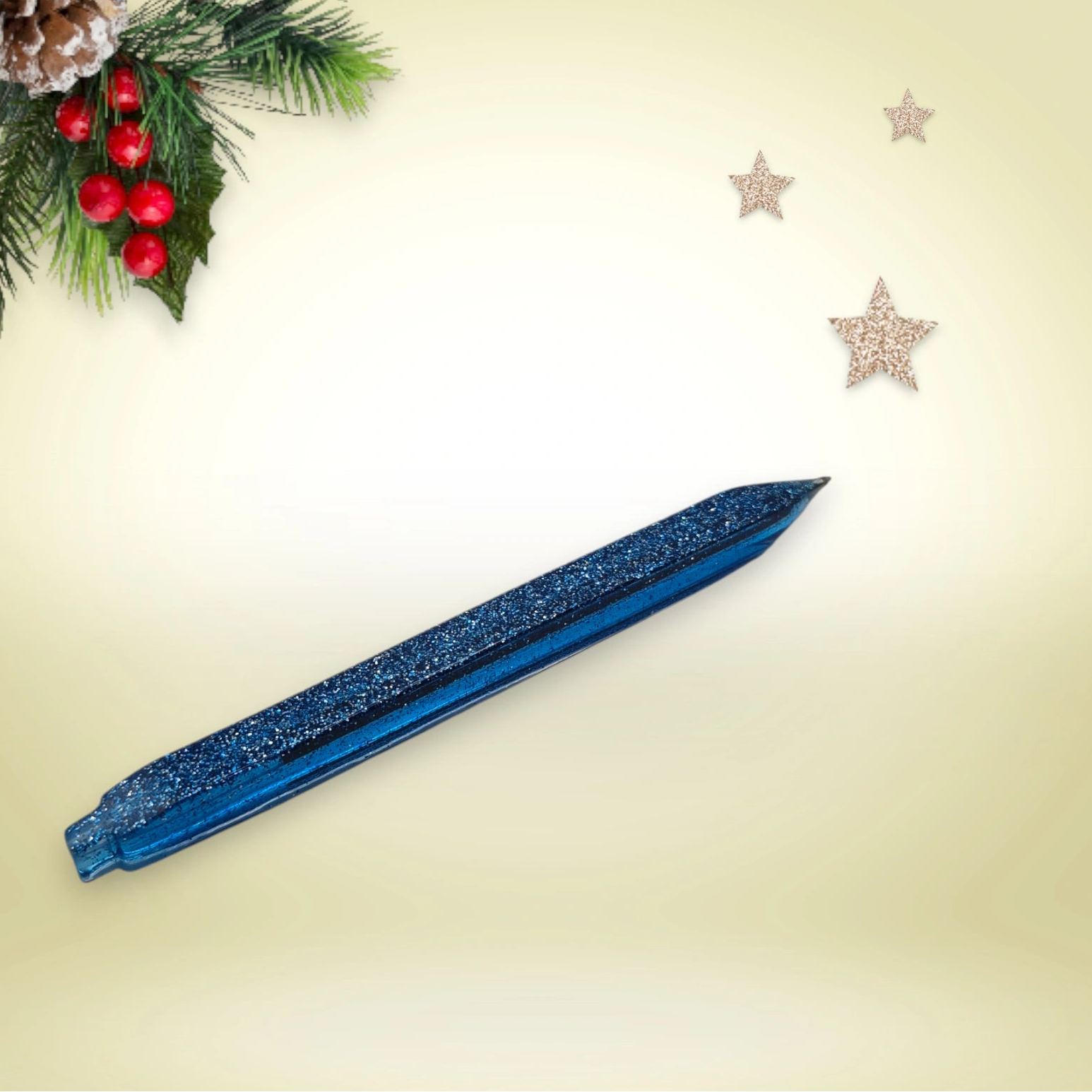 Stylo bleu pailleté en résine époxy avec encre noire, présenté sur un décor de Noël composé de branches de pin et d’étoiles festives.