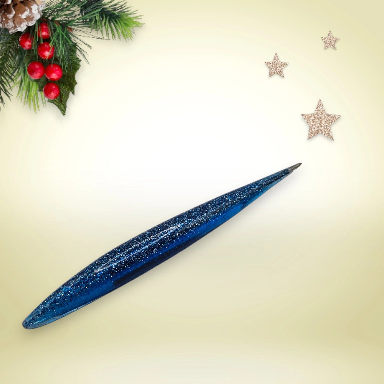 Stylo bleu pailleté en résine époxy avec encre noire, présenté sur un décor de Noël composé de branches de pin et d’étoiles festives.