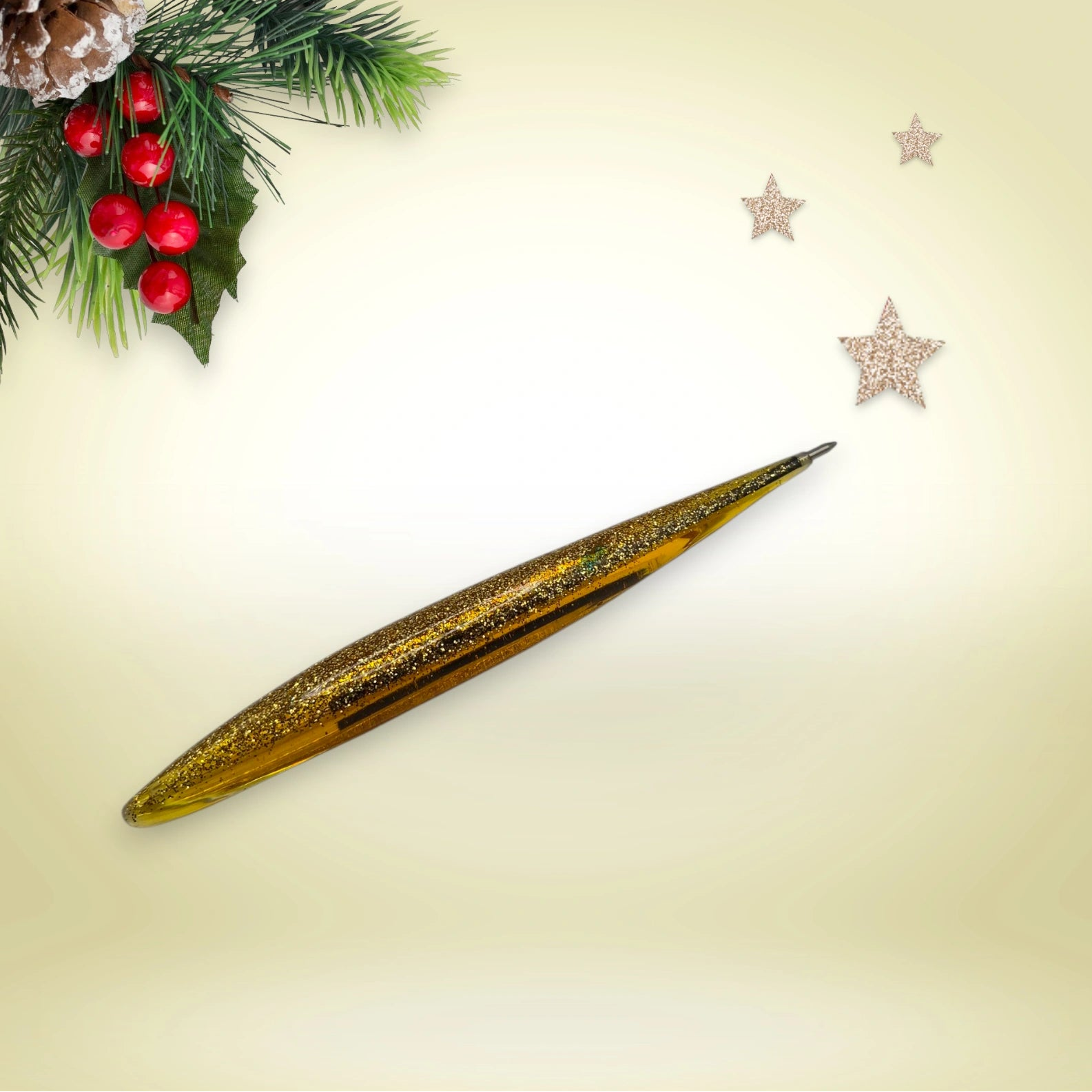 Stylo doré pailleté en résine époxy avec encre noire, présenté sur un décor de Noël composé de branches de pin et d’étoiles festives.