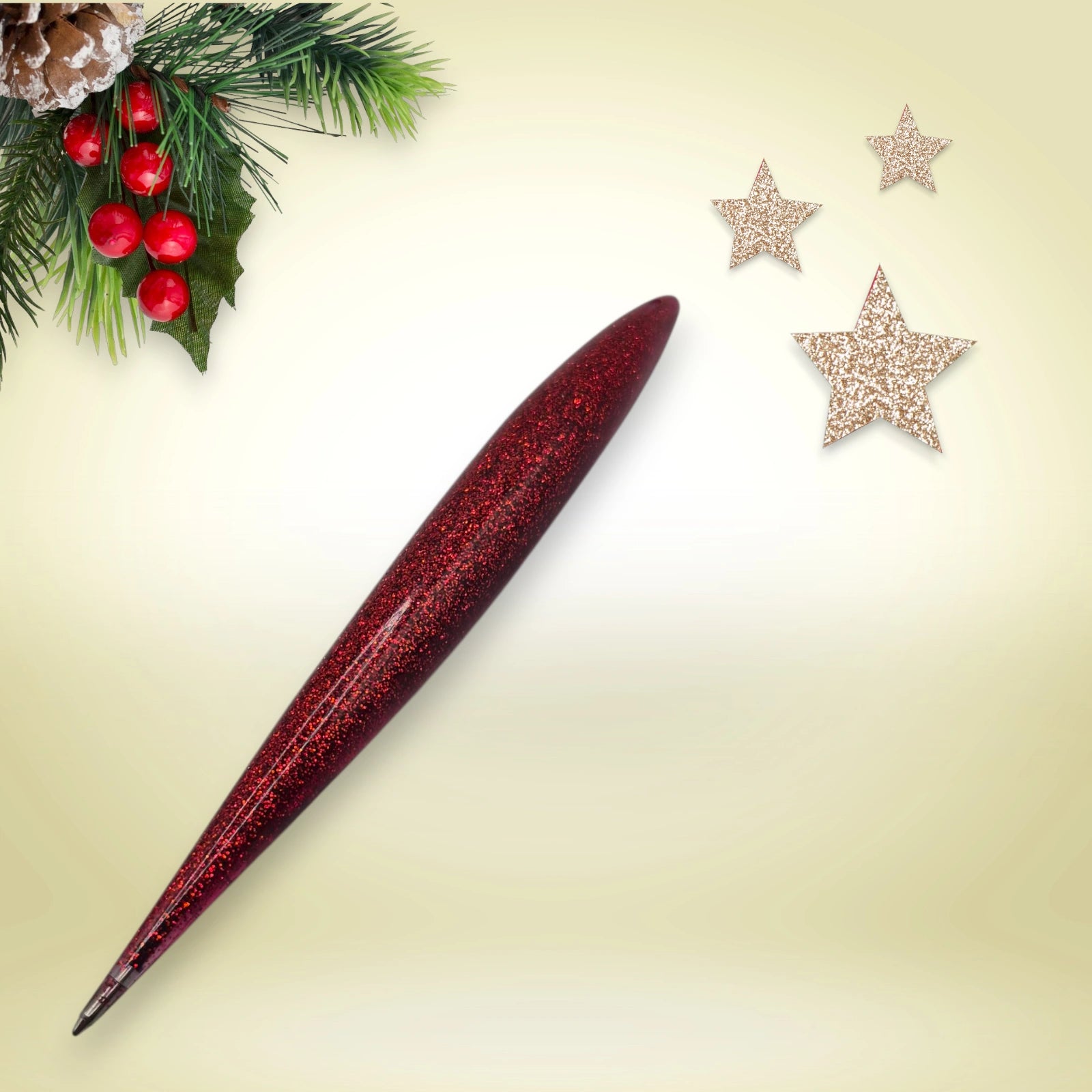 Stylo rouge pailleté en résine époxy avec encre noire, présenté sur un décor de Noël composé de branches de pin et d’étoiles festives.