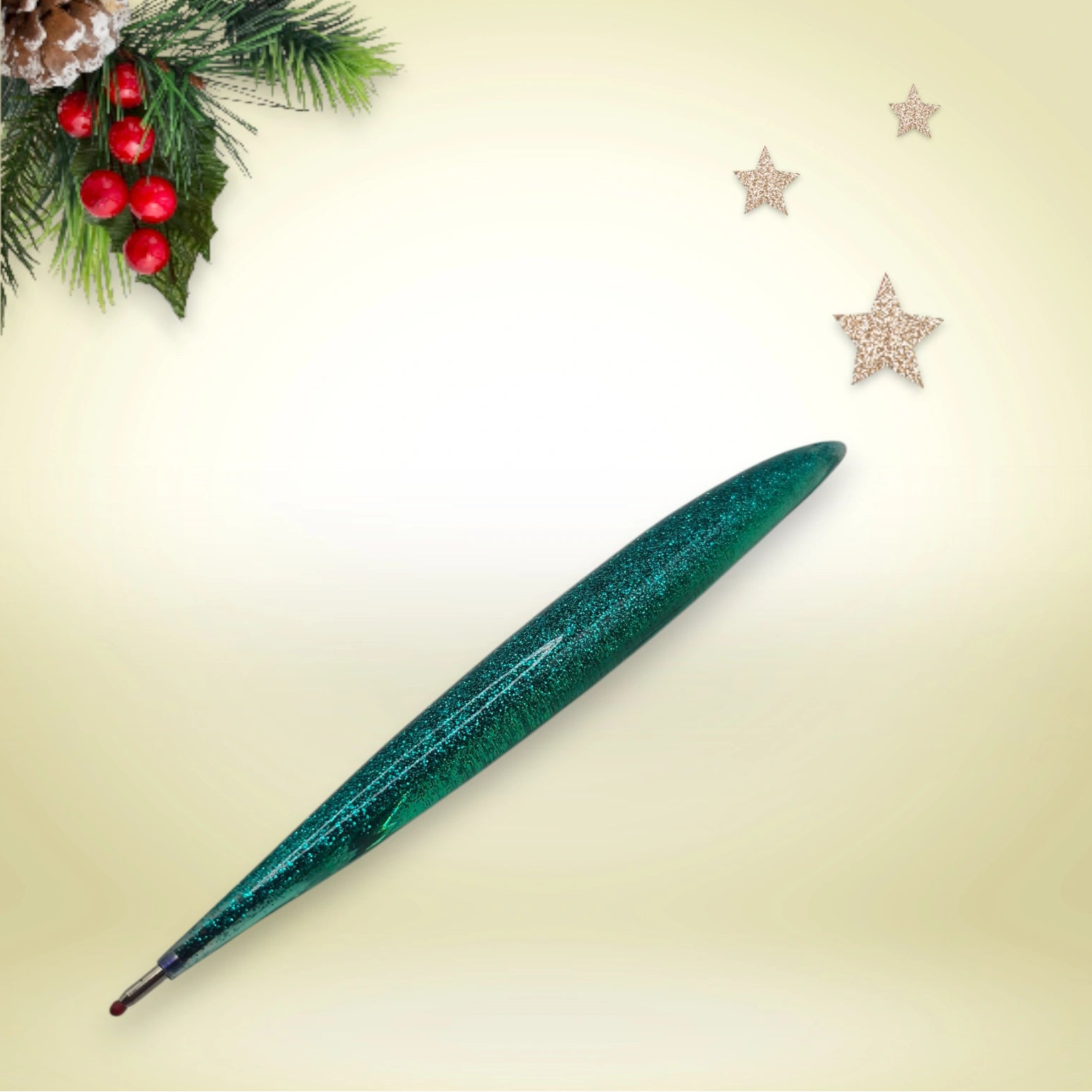 Stylo vert pailleté en résine époxy avec encre noire, présenté sur un décor de Noël composé de branches de pin et d’étoiles festives.