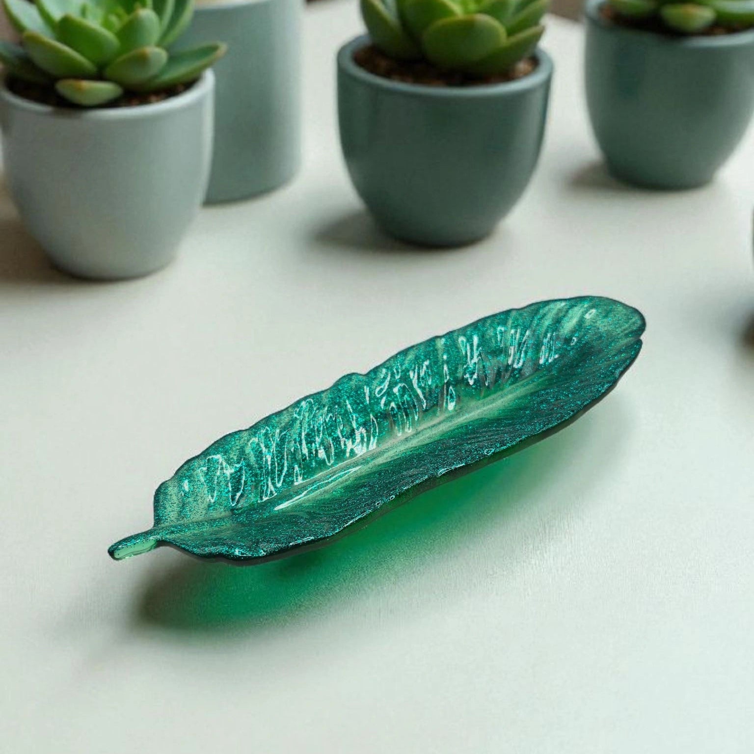 vide-poche en forme de plume en résine pailletée verte posée sur une surface plane avec en arrière-plan des petits pots de plantes.